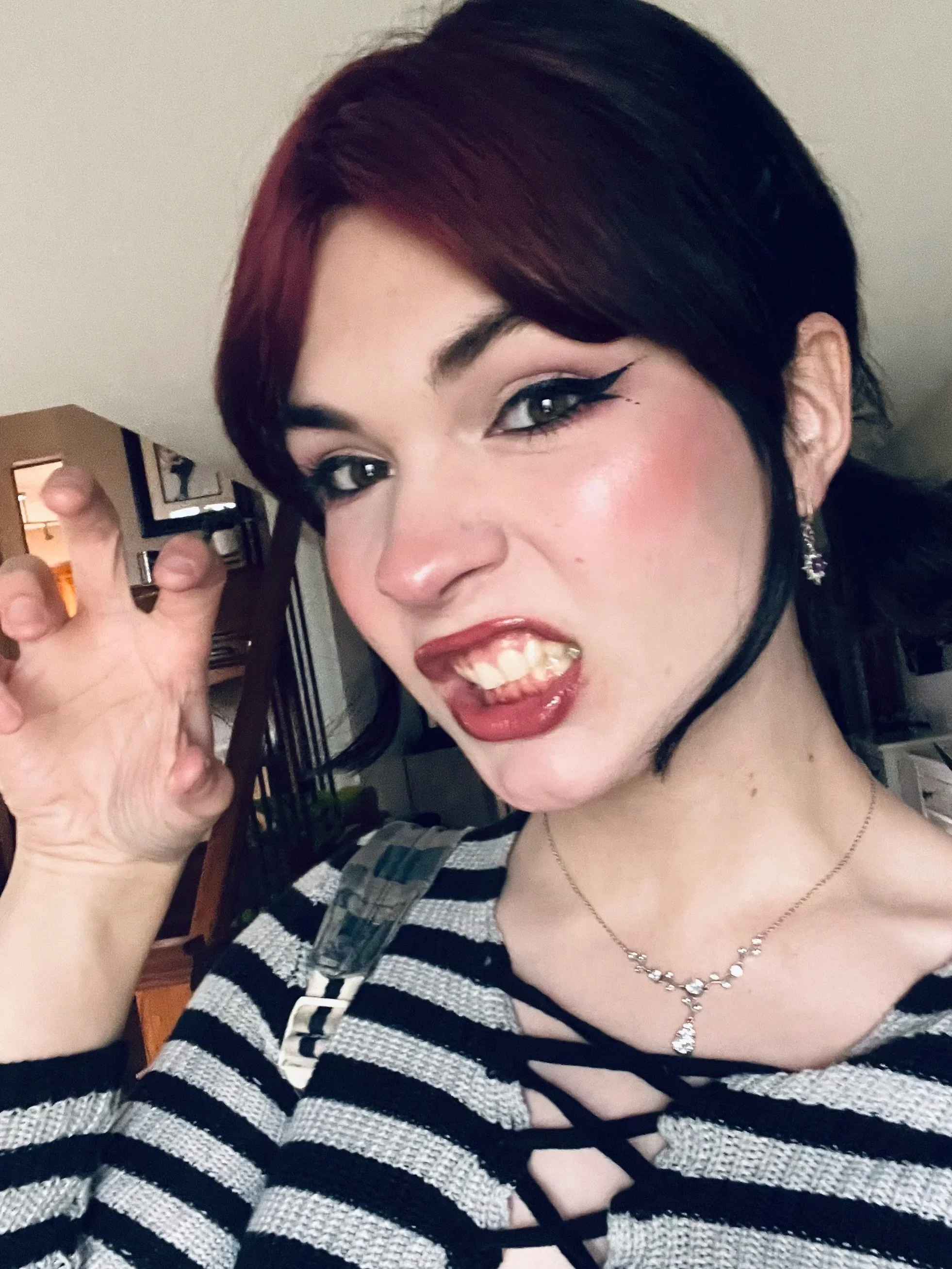 A young woman with dark, short hair styled to the side, wearing a striped top, a necklace, and earrings. She has makeup with winged eyeliner and is smiling with her teeth showing, making a claw gesture.