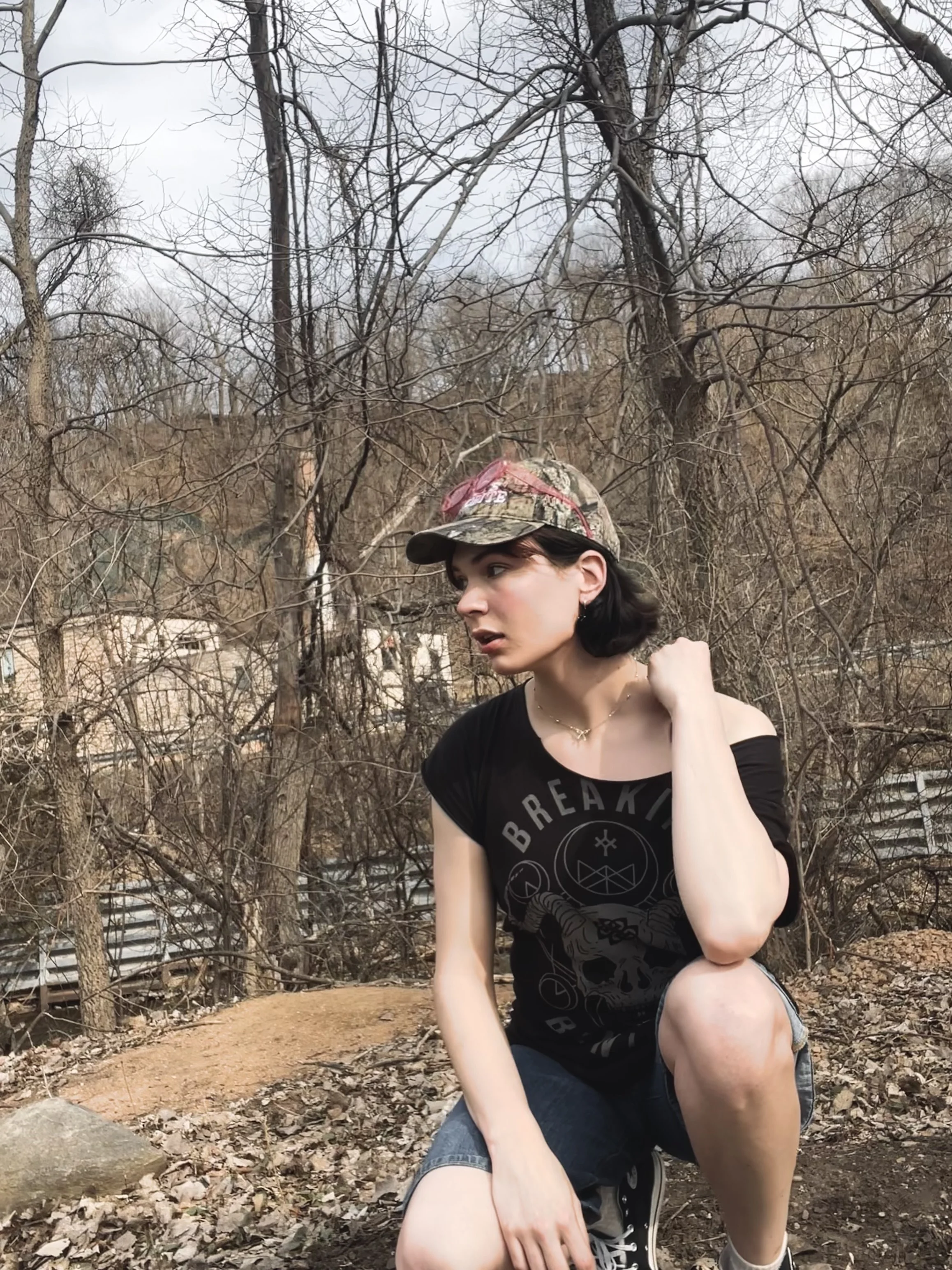 A woman with dark hair, wearing a camouflage cap and black T-shirt, crouches outdoors in a wooded area with leafless trees and a fence in the background.