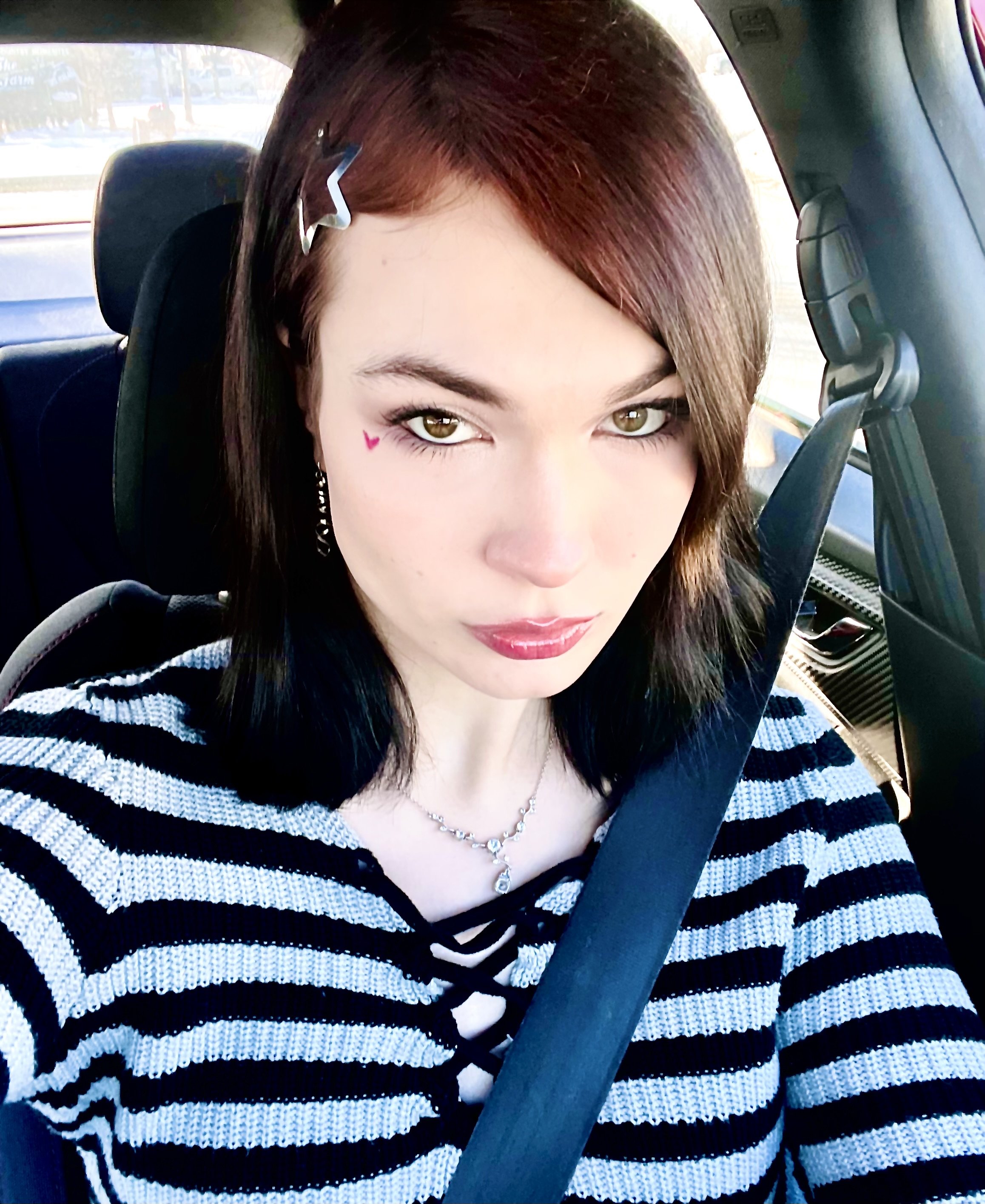 A woman with brown hair and hazel eyes wearing a black and white striped sweater, a necklace, and a heart-shaped face sticker, sitting in a car and wearing a seatbelt.