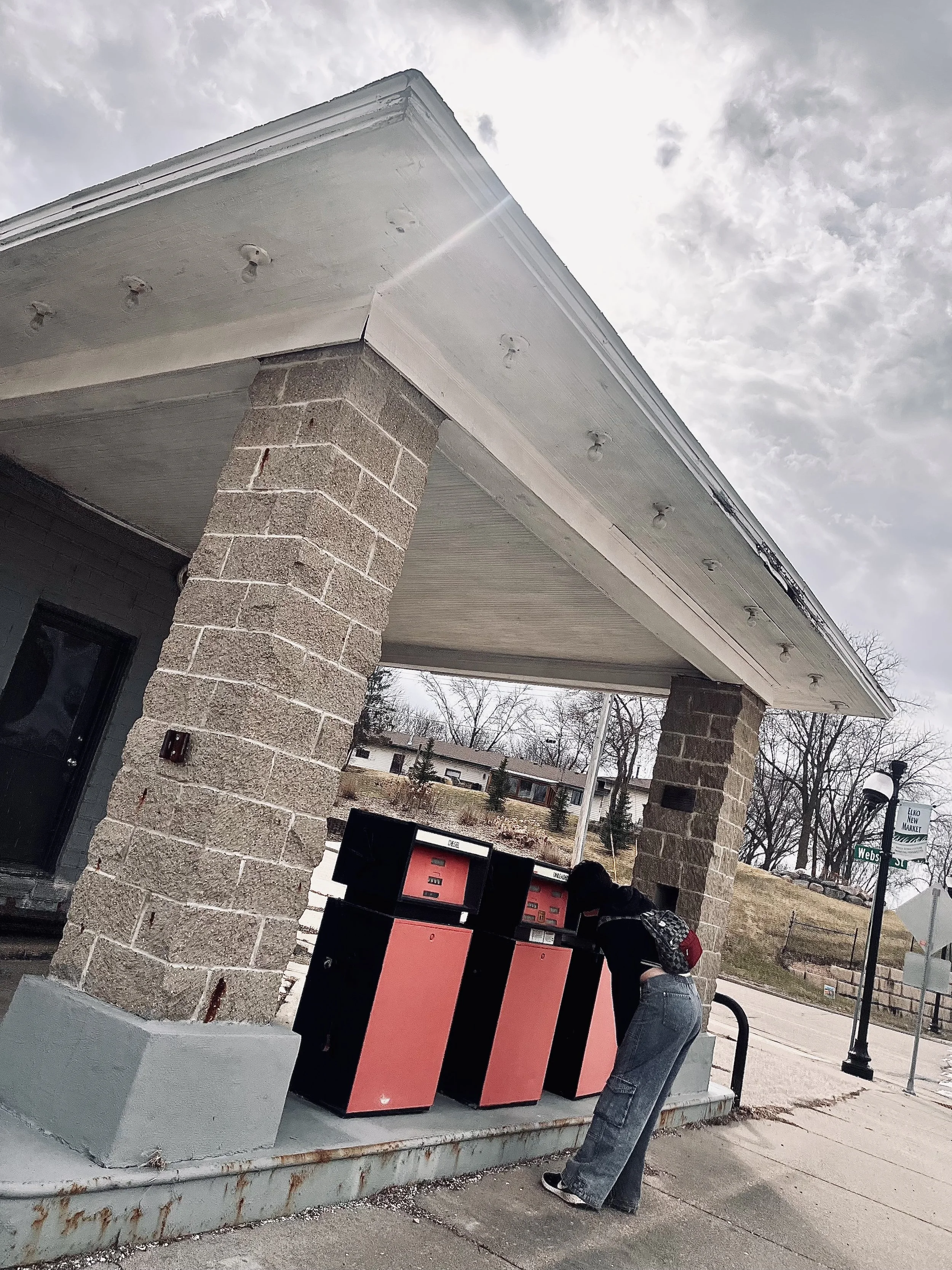 A person in a dark jacket and wide-legged jeans leaning against a large, pink and black postal mail drop box at a sidewalk. The building has brick pillars and a white roof with external light fixtures, and the sky is cloudy.