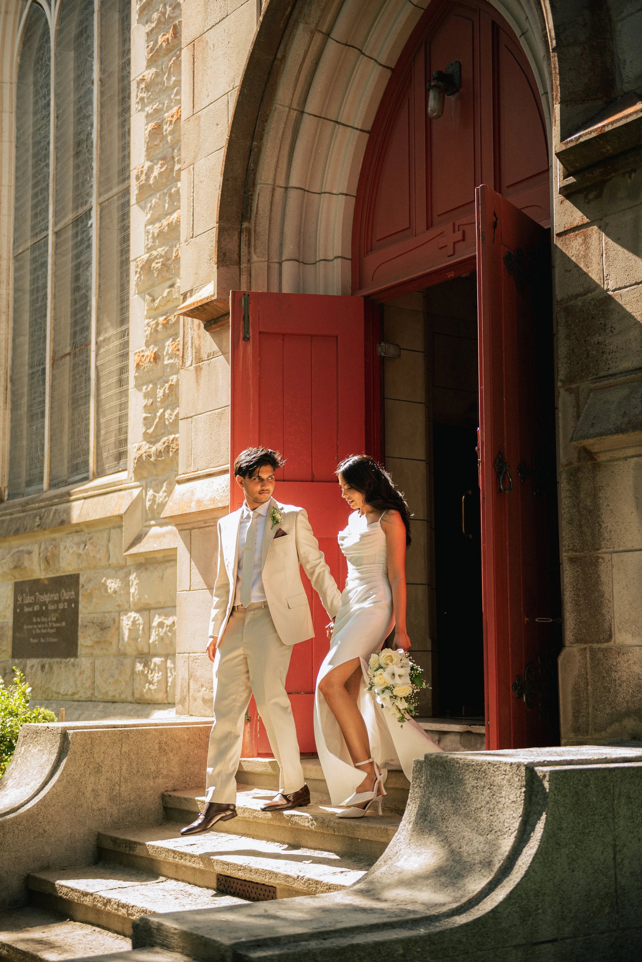 A couple in wedding attire exiting a church with red doors, holding hands and a flower bouquet.