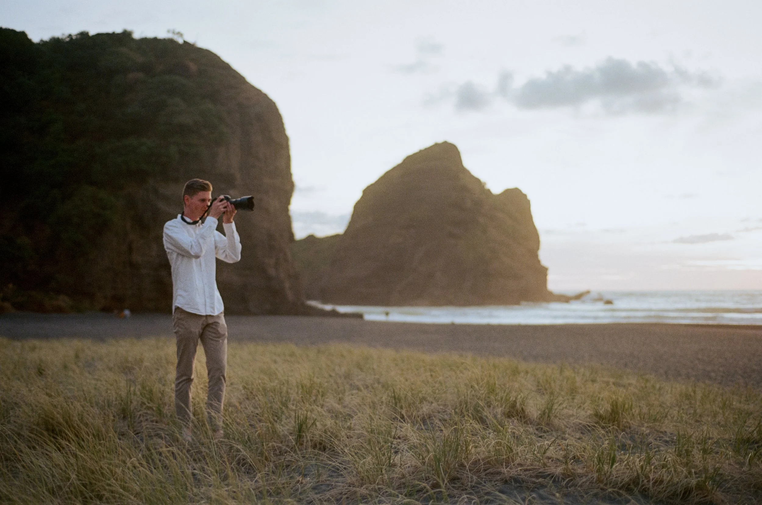A man taking a photo with a camera on a beach with large rocky cliffs and the ocean in the background at sunset.