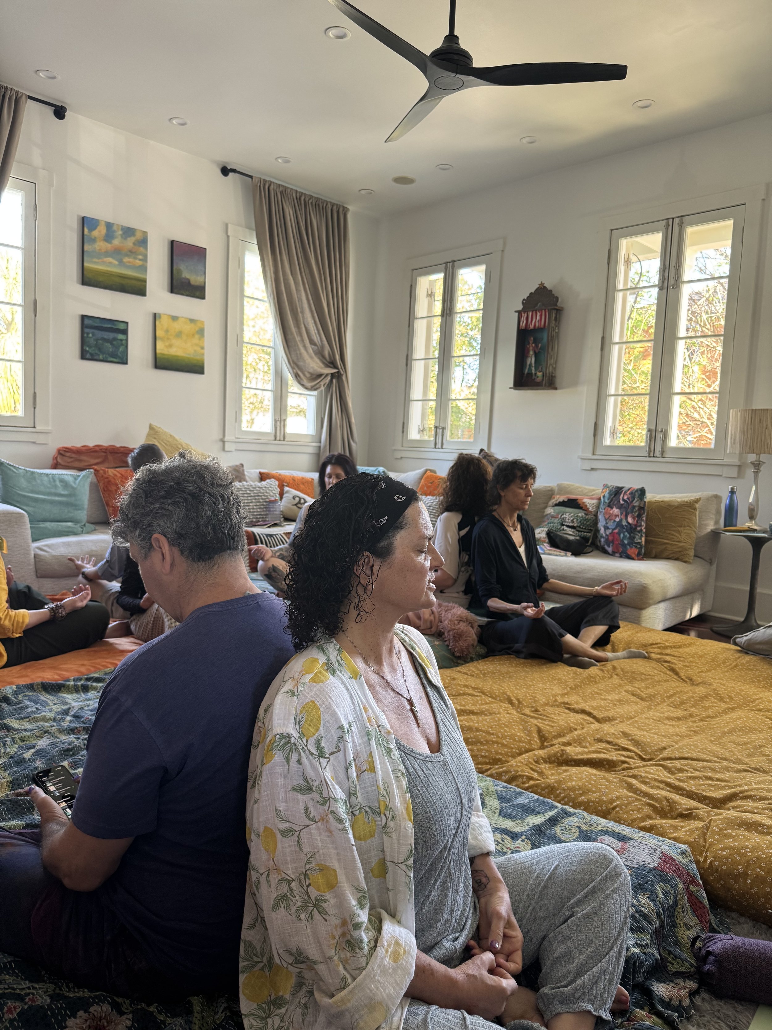 People sitting cross-legged on the floor in a brightly lit room with large windows, curtains, and colorful pillows, participating in a somatic exercise during a creative workshop at Seducing the Muse retreat.