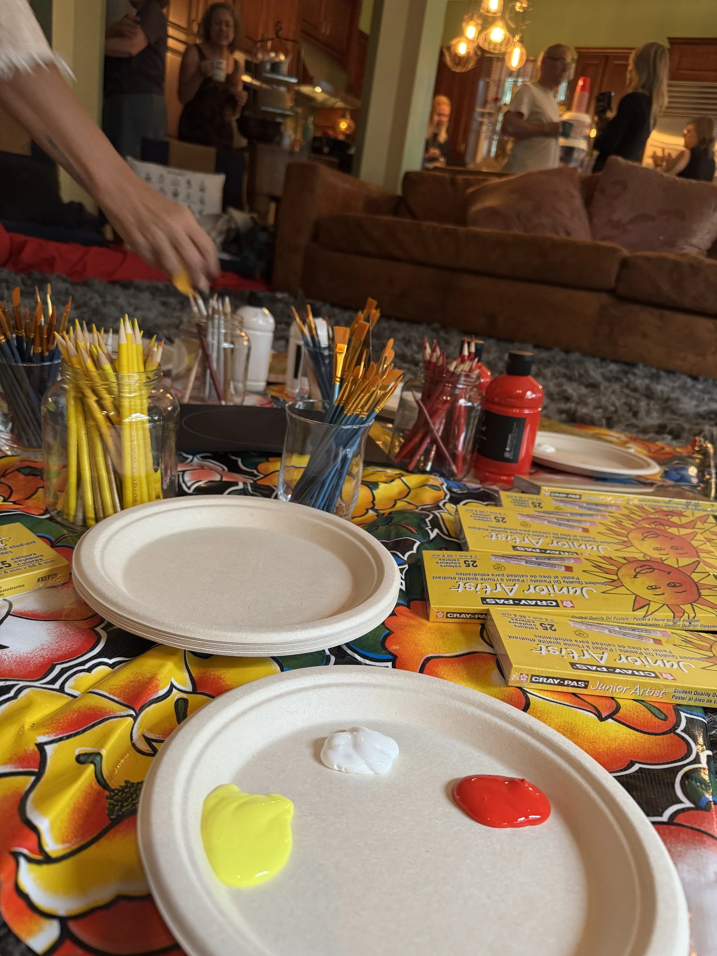 A table with a colorful tablecloth, art supplies including yellow and red oil pastels, and a paper plate with dabs of yellow, white, and red paint, in a living room with people socializing.