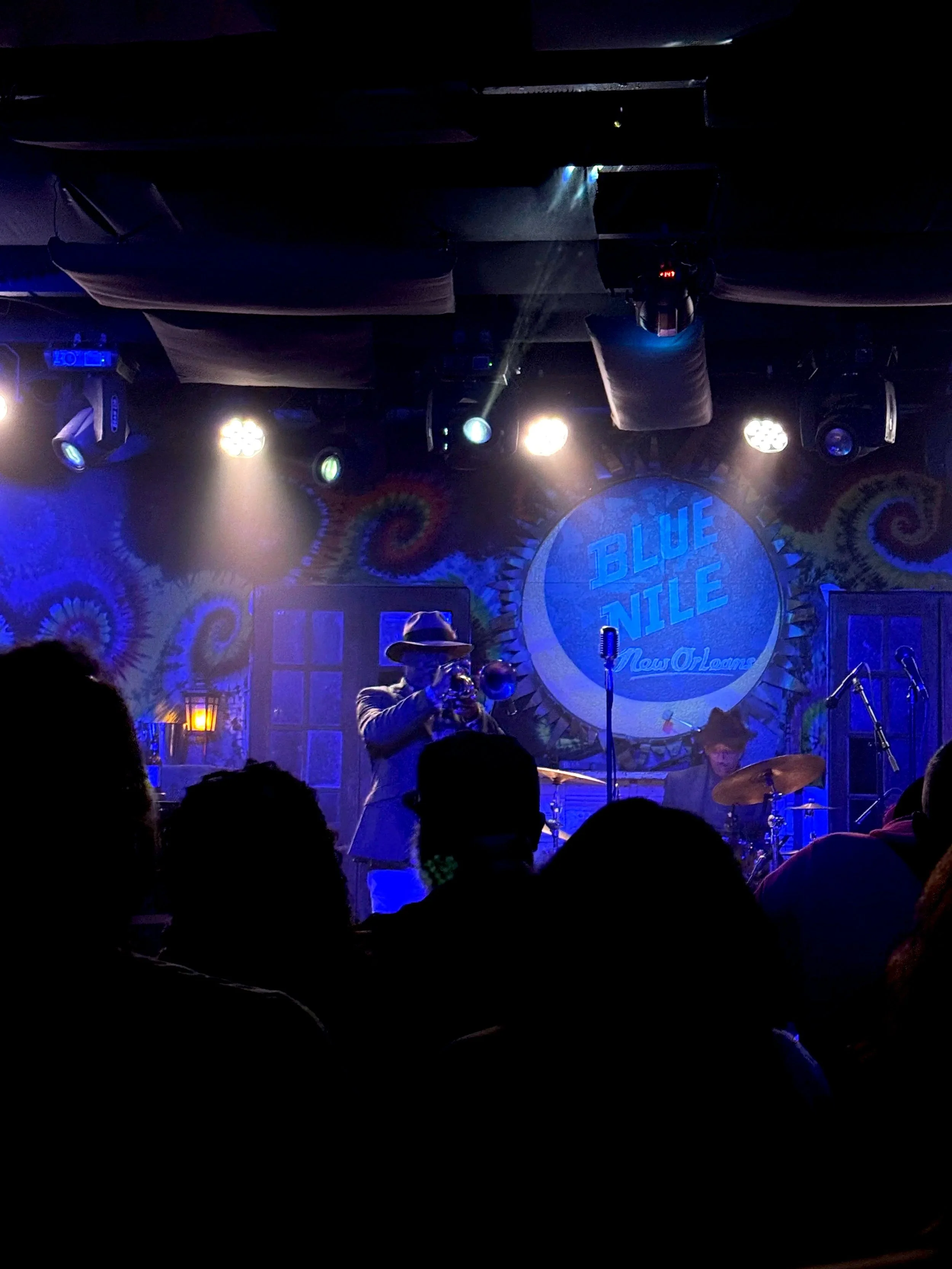 A live music performance at Blue Nile in New Orleans, featuring a trumpet player in a cowboy hat and a drummer on stage surrounded by colorful stage lighting.