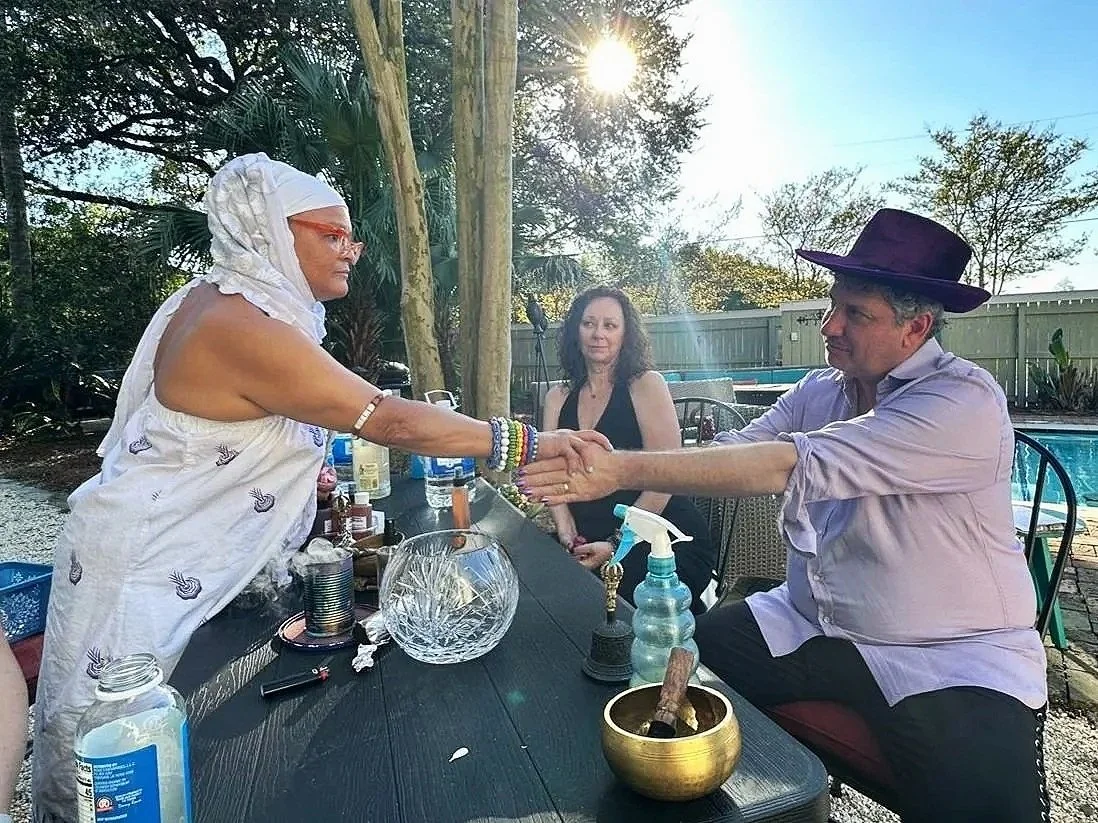 A Yoruba blessing ritual at Seducing the Muse in New Orleans. The man on the right is wearing a large purple hat, and the woman on the left is wearing glasses and a white headscarf.