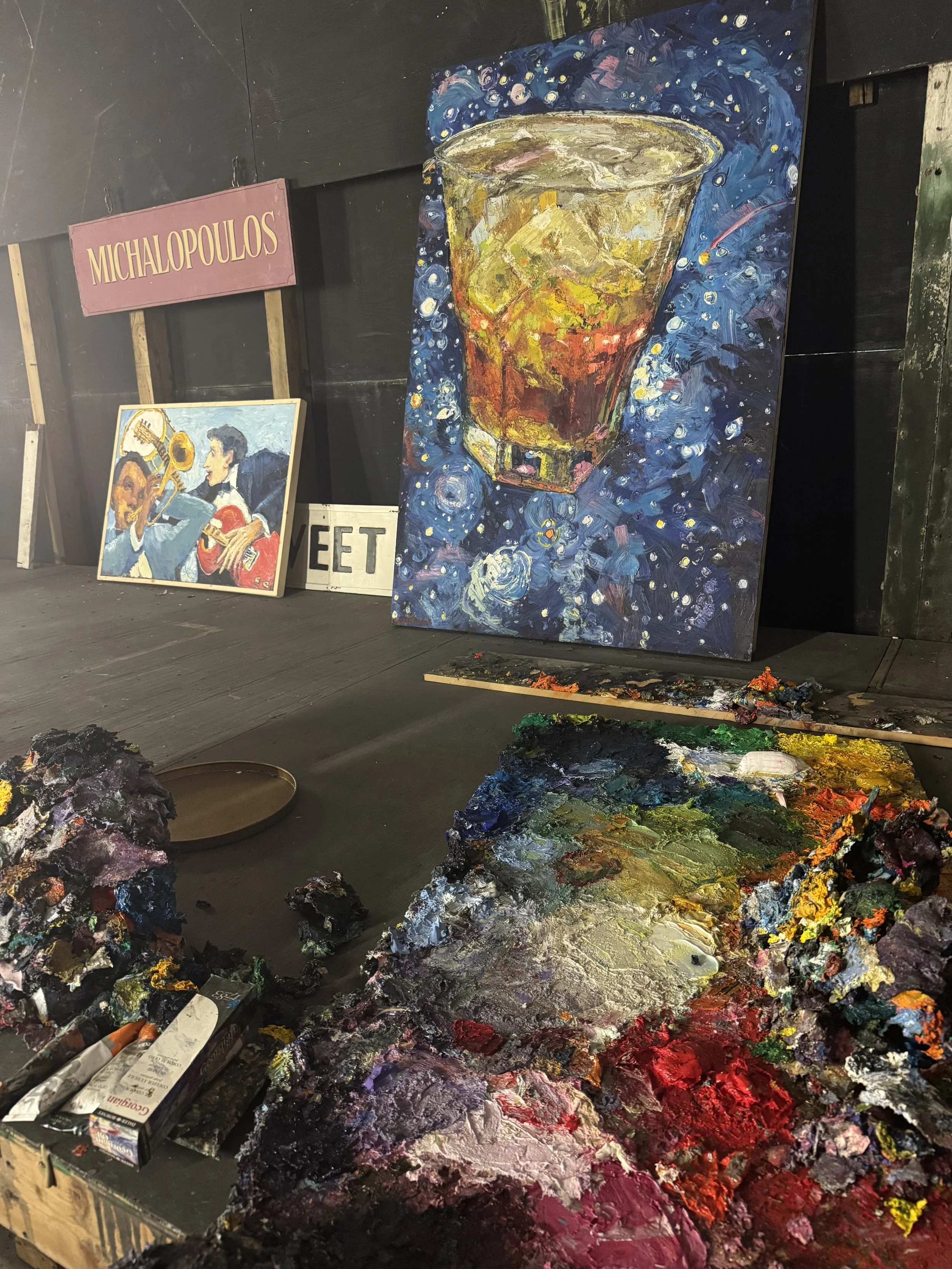 Colorful acrylic painting of a glass of whiskey with ice cubes on a dark blue, starry background, alongside palettes of thick, textured paint at the studio of artist James Michalopoulos in New Orleans. 