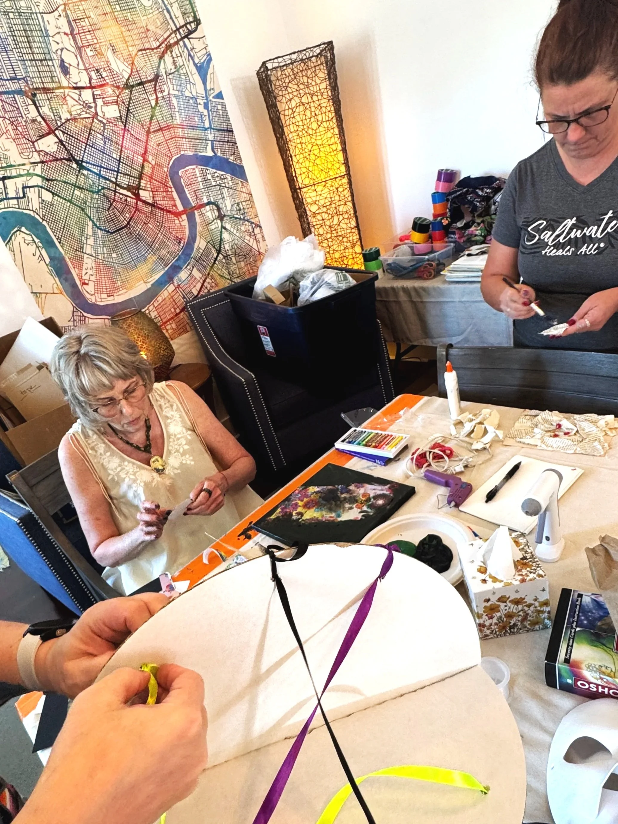 People working on arts and crafts projects at a table during Seducing the Muse, with various art supplies, a large colorful map on the wall, and a tall decorative lamp in the background.