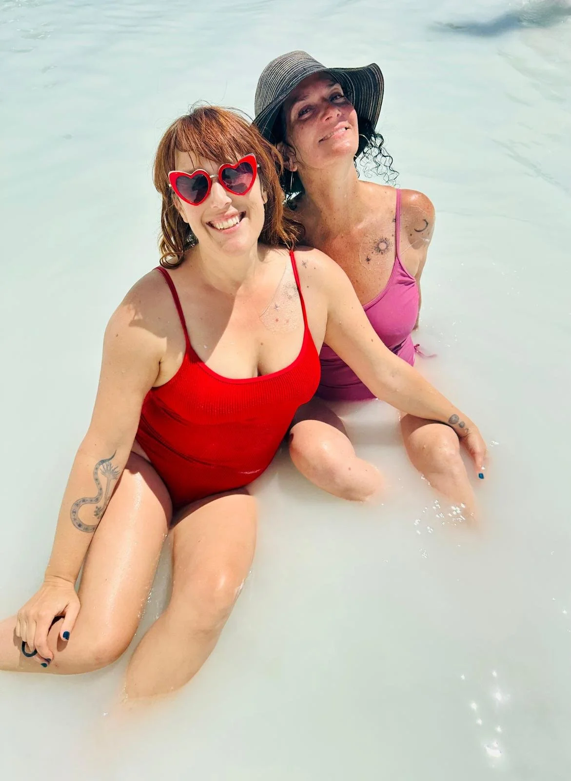 The facilitators of Seducing the Muse, Lara Blackin and Joanna Garner, sitting in milky-white water, smiling at the camera. One woman wears a red swimsuit and heart-shaped sunglasses, while the other wears a pink swimsuit and a large sun hat.