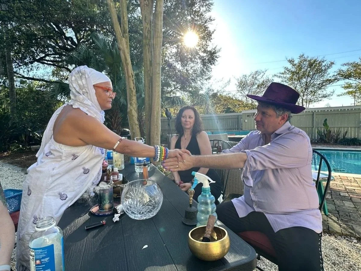 A Yoruba blessing ritual at Seducing the Muse in New Orleans. The man on the right is wearing a large purple hat, and the woman on the left is wearing glasses and a white headscarf.