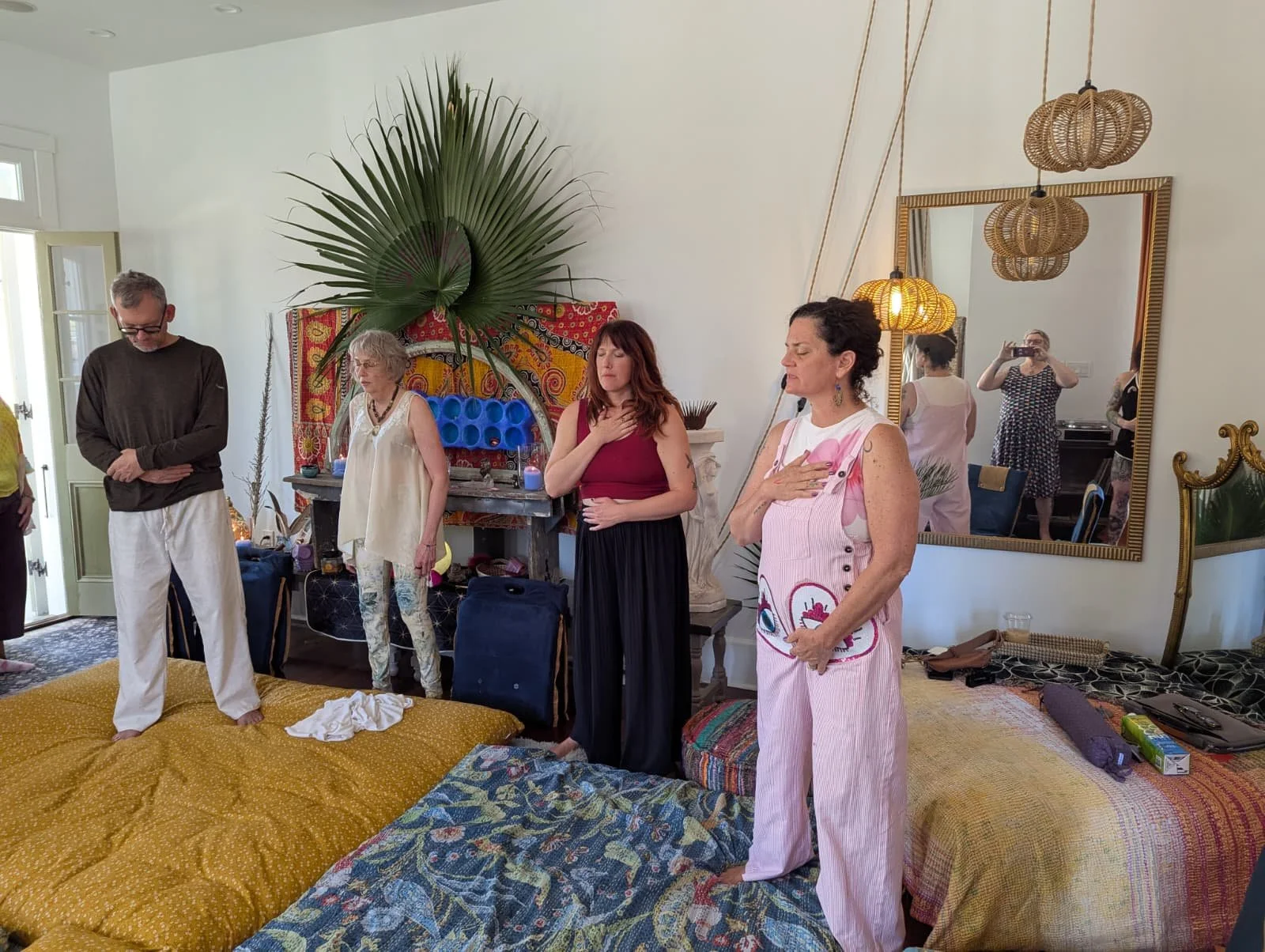 People standing with their hands over their hearts in a room with vibrant decorations, a large mirror, and layered mattresses on the floor during the creative workshop at Seducing the Muse retreat.