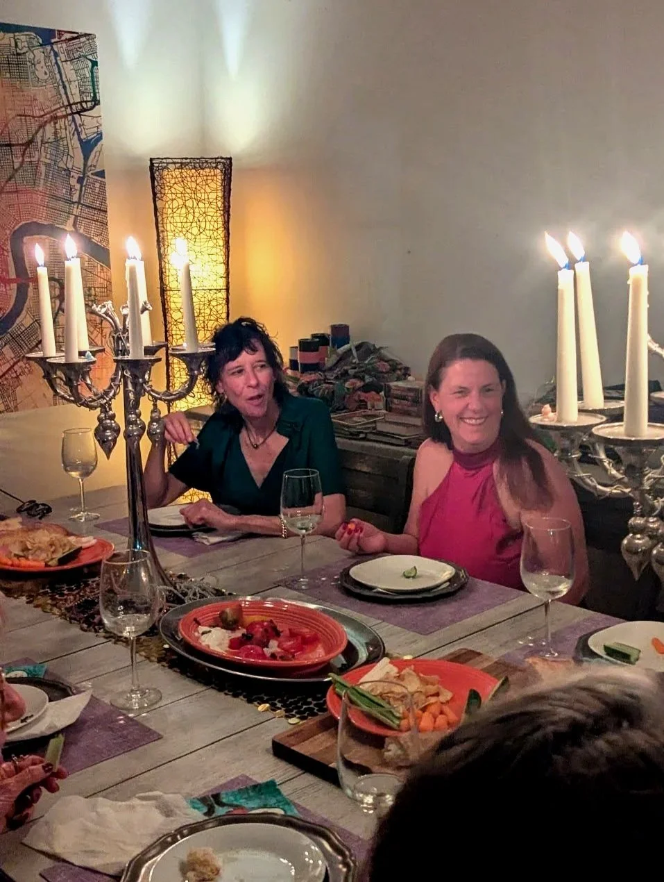 Two women sitting at a dining table during a dinner party at Seducing the Muse, with lit white candles in ornate candelabras, plates of food, wine glasses, and a large map on the wall in the background.