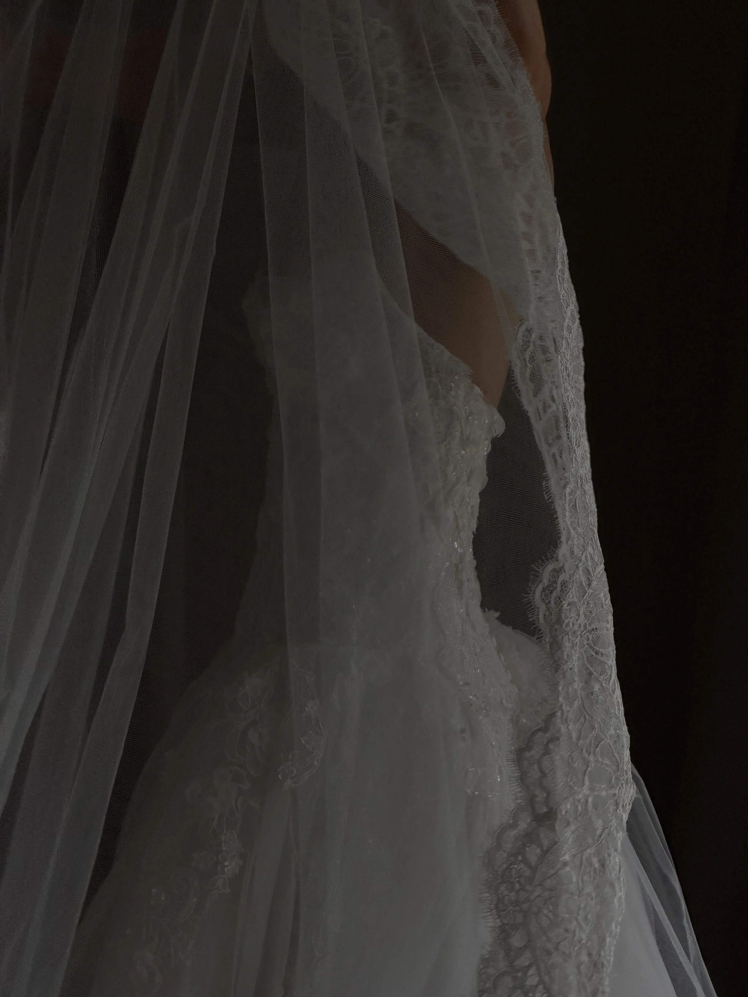 Close-up of a sheer white lace and tulle wedding veil with intricate lace trim, displayed on a dark surface.