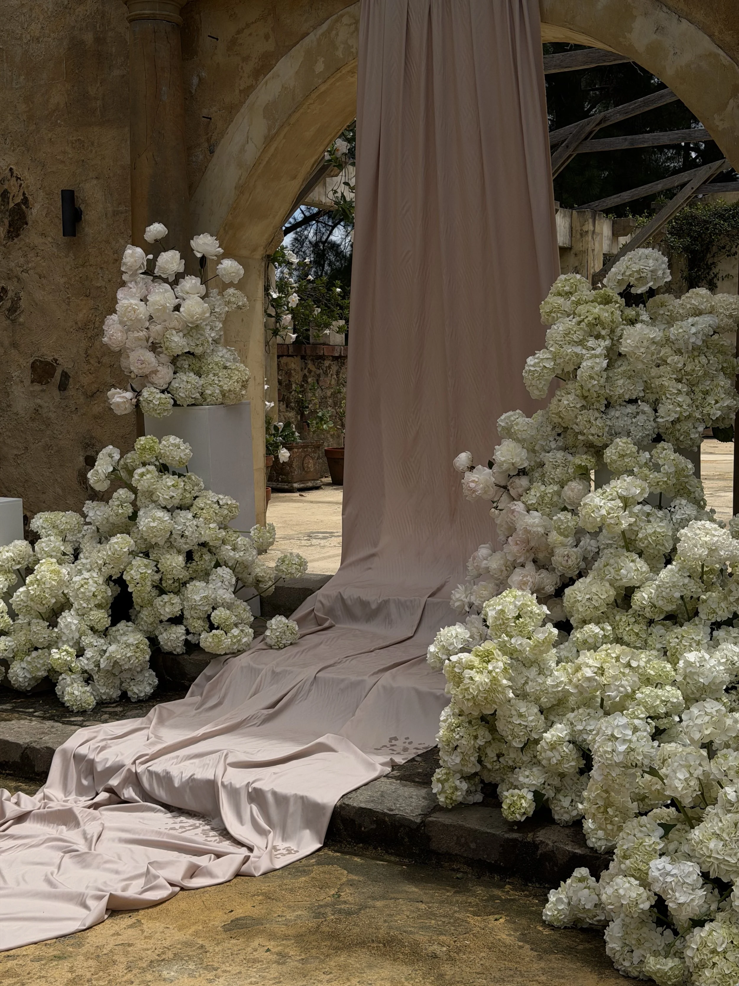 A decorative outdoor setting with white hydrangea flowers, a pink fabric backdrop, and an archway made of stone and wood.
