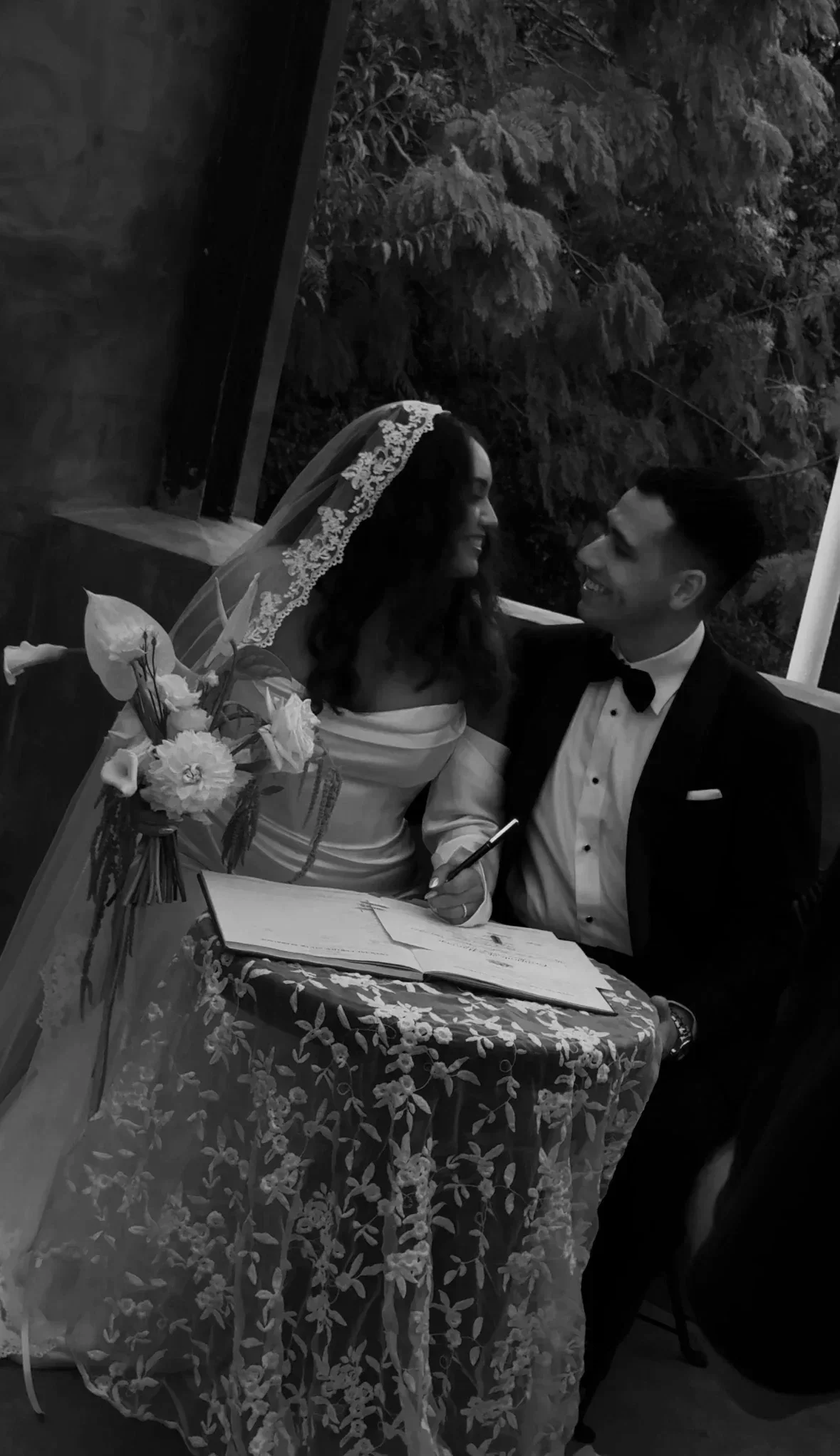 A bride and groom smiling at each other in a black and white photo, sitting at a table with a floral tablecloth, with trees visible outside the window.