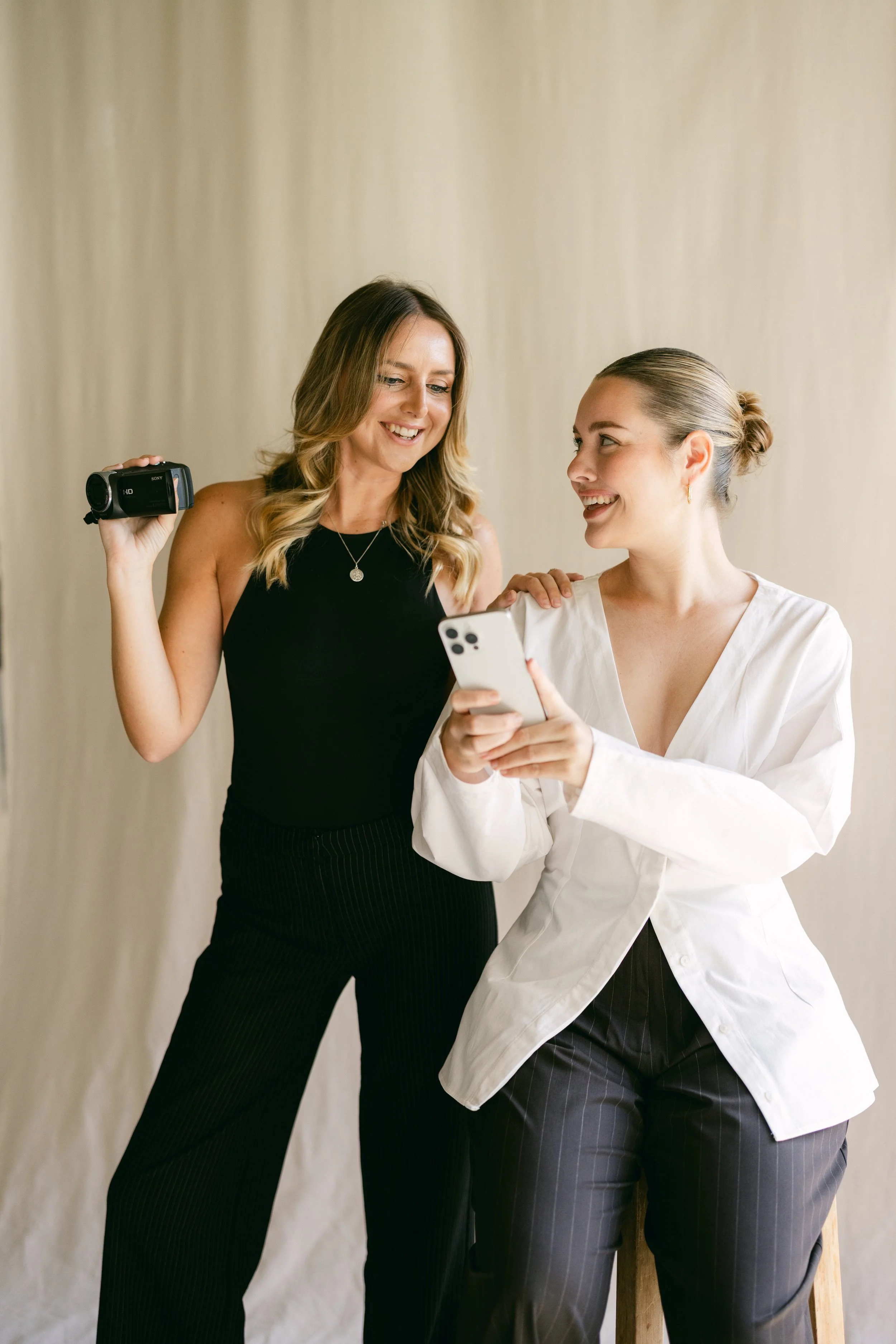 Two women smiling, one holding a video camera and the other looking at her phone, standing against a neutral background.
