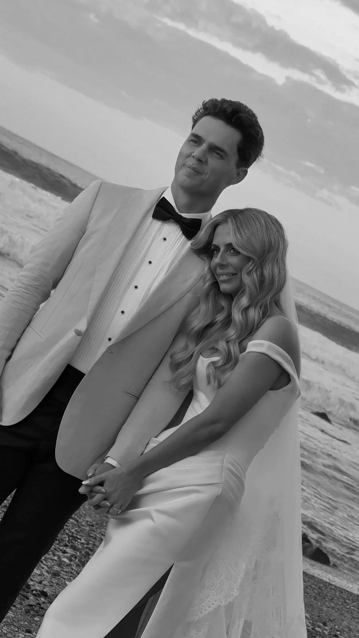 A black and white photo of a man and woman on the beach, dressed in formal wedding attire, standing close together, smiling, with the ocean and sky in the background.