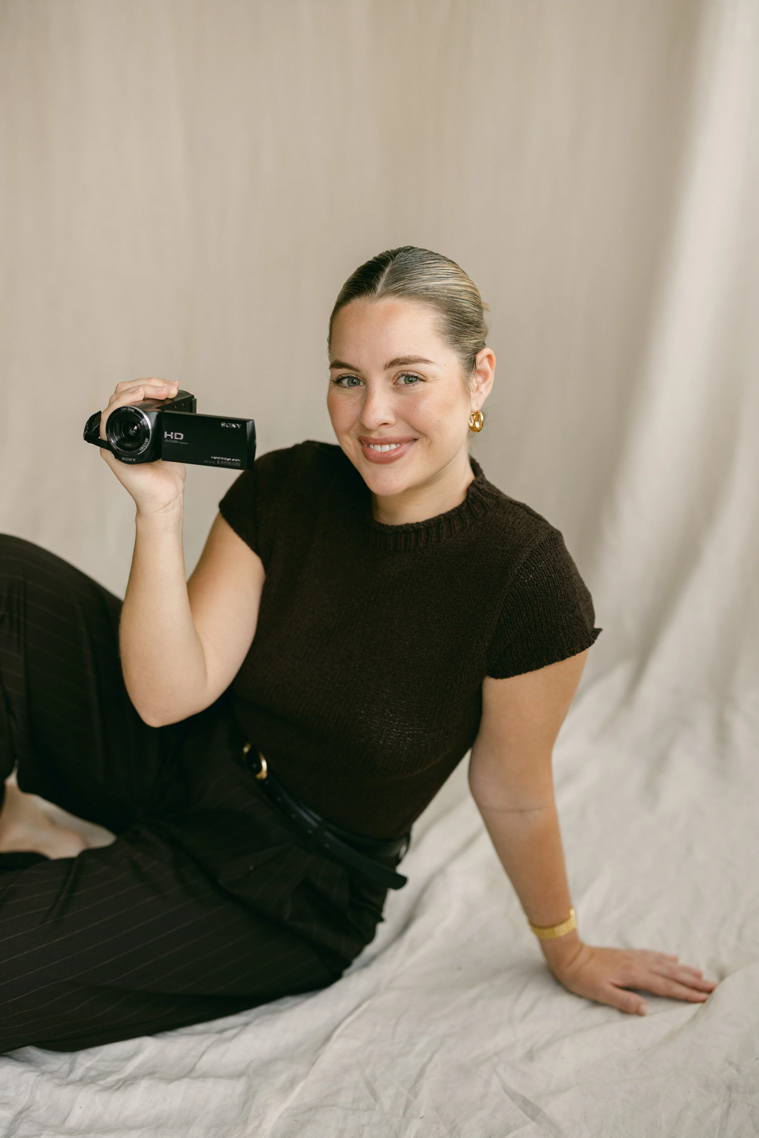 A smiling woman with brown hair slicked back, wearing gold earrings, a dark brown sweater, and black striped pants, sitting on a white sheet, holding a small black Sony HD camcorder, with a beige background.