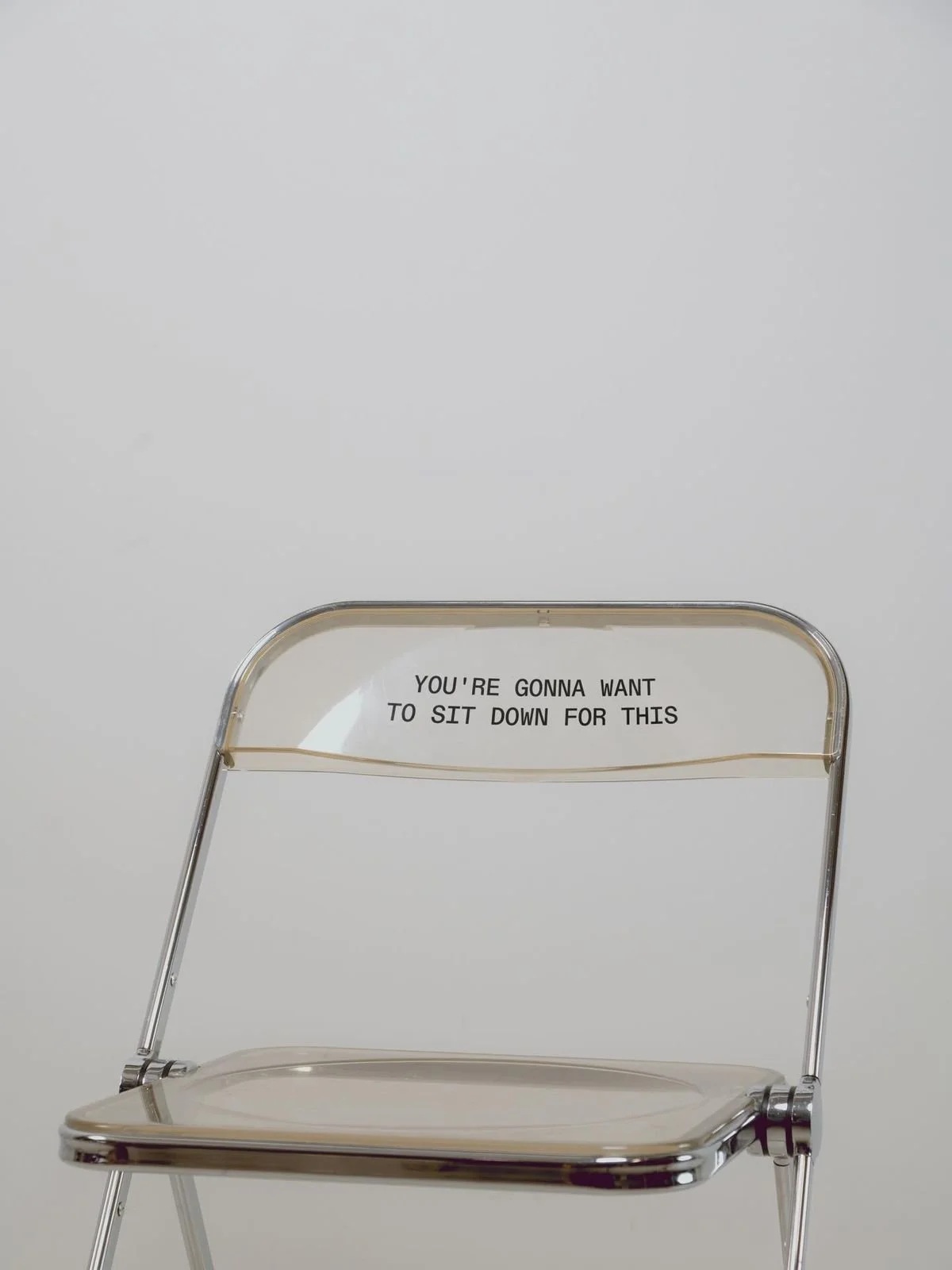 A clear plastic chair with a message on the backrest that reads, 'You're gonna want to sit down for this.'