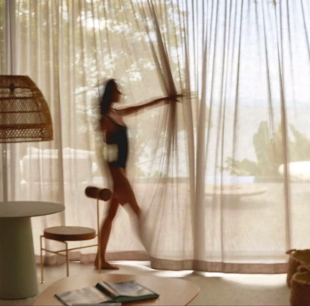 A person in a black bikini standing by sheer curtains, pulling them aside, with sunlight coming through and trees visible outside.