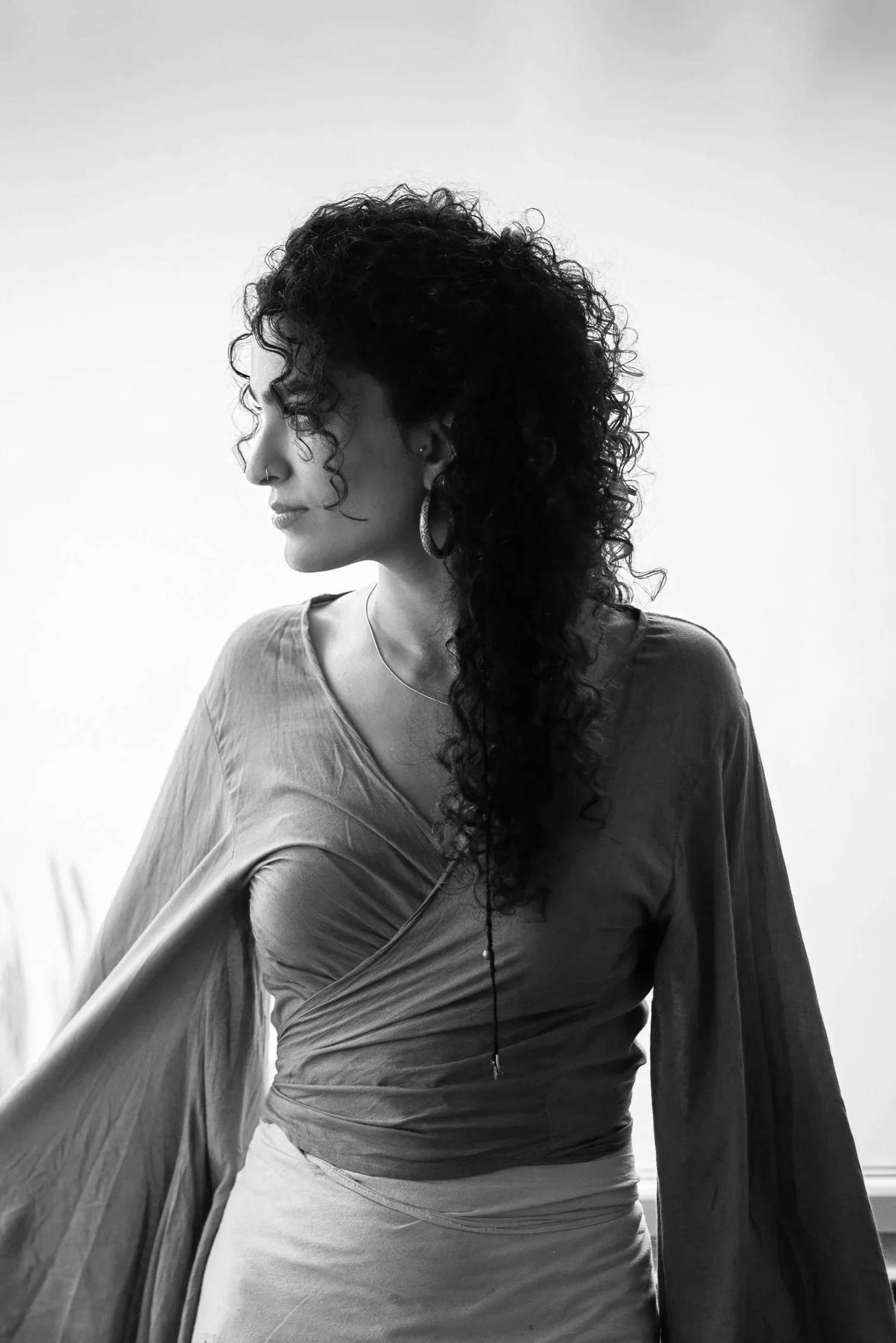 Black and white photo of a woman with curly hair looking to her left, wearing a draped top, earrings, and a nose ring, standing against a plain background.