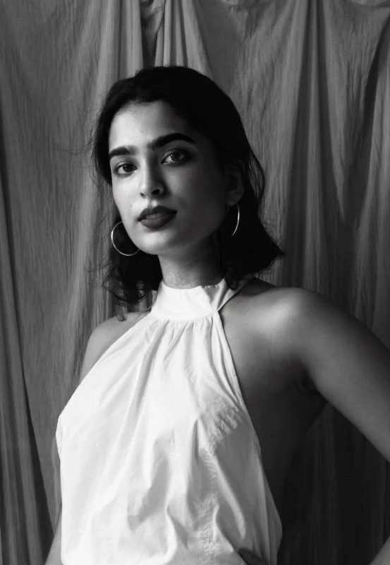 Black and white photo of a woman with dark hair wearing hoop earrings and a sleeveless top, standing in front of curtains.