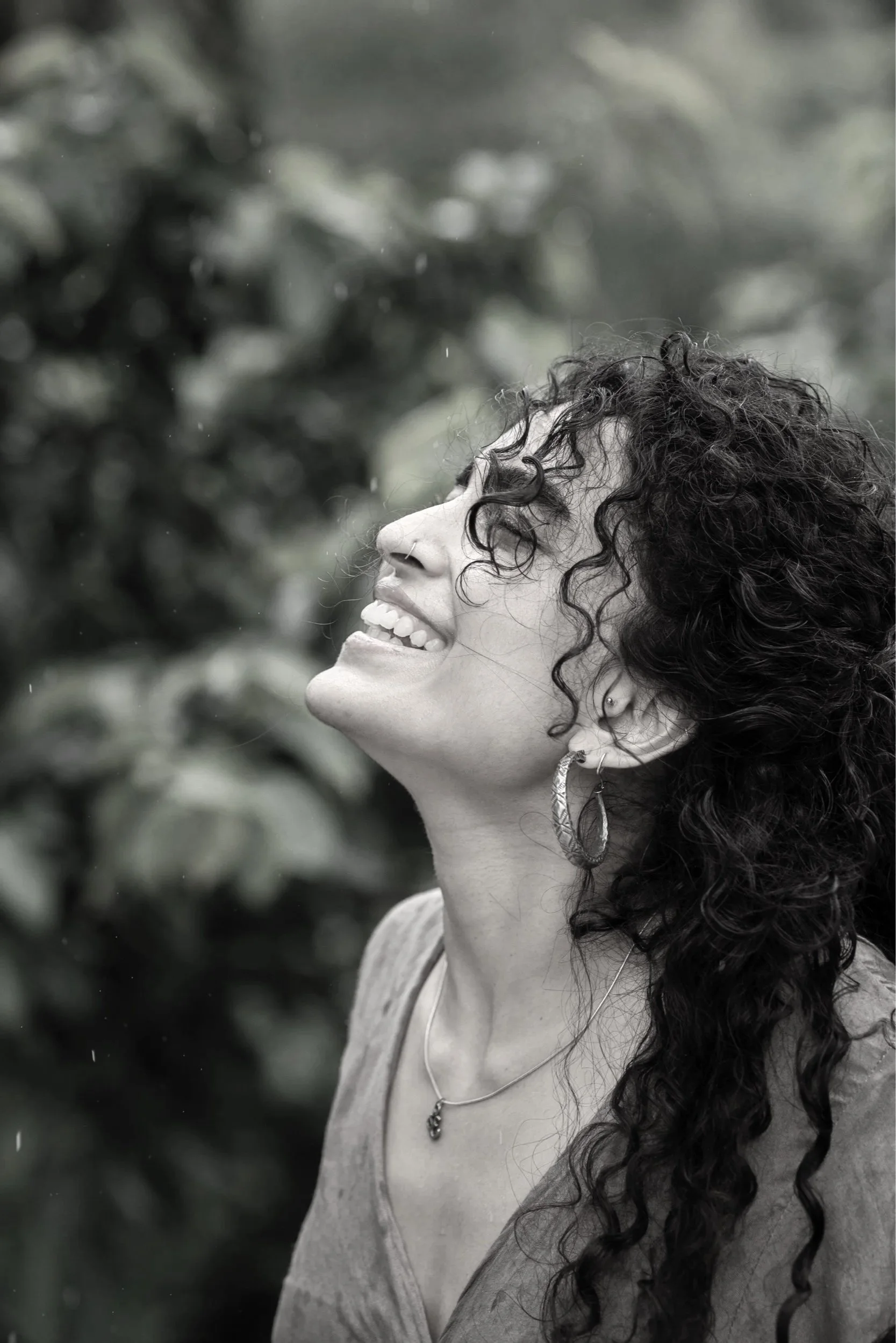 A woman with curly dark hair smiling and looking upwards, wearing earrings, a necklace, and a sleeveless top, outdoors with blurred foliage in the background.