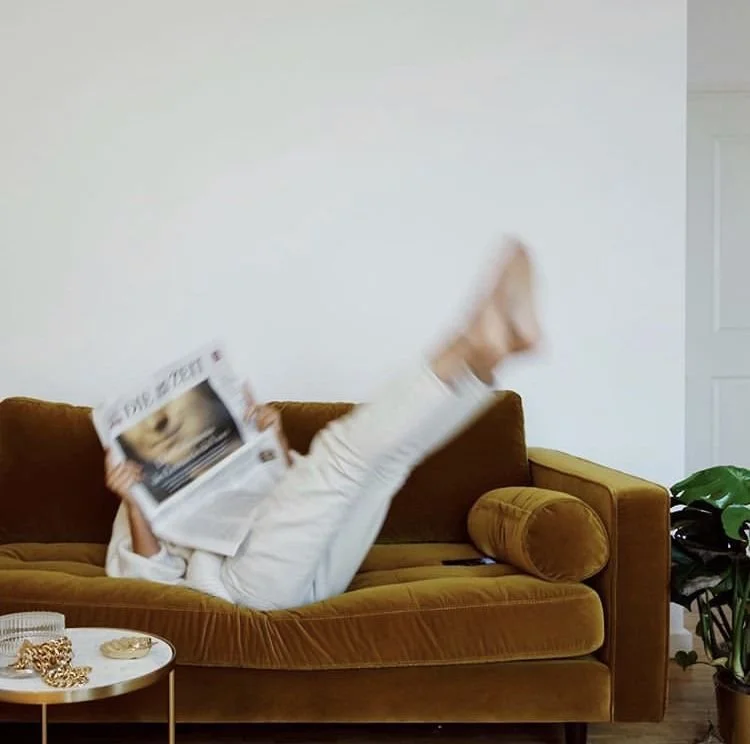 Person lying on a brown velvet sofa with legs raised, reading a newspaper.