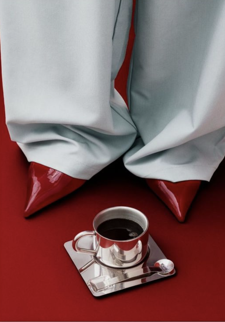 A stainless steel cup of black coffee on a reflective silver tray, resting on a red surface with crumpled white and gray fabric in the background.