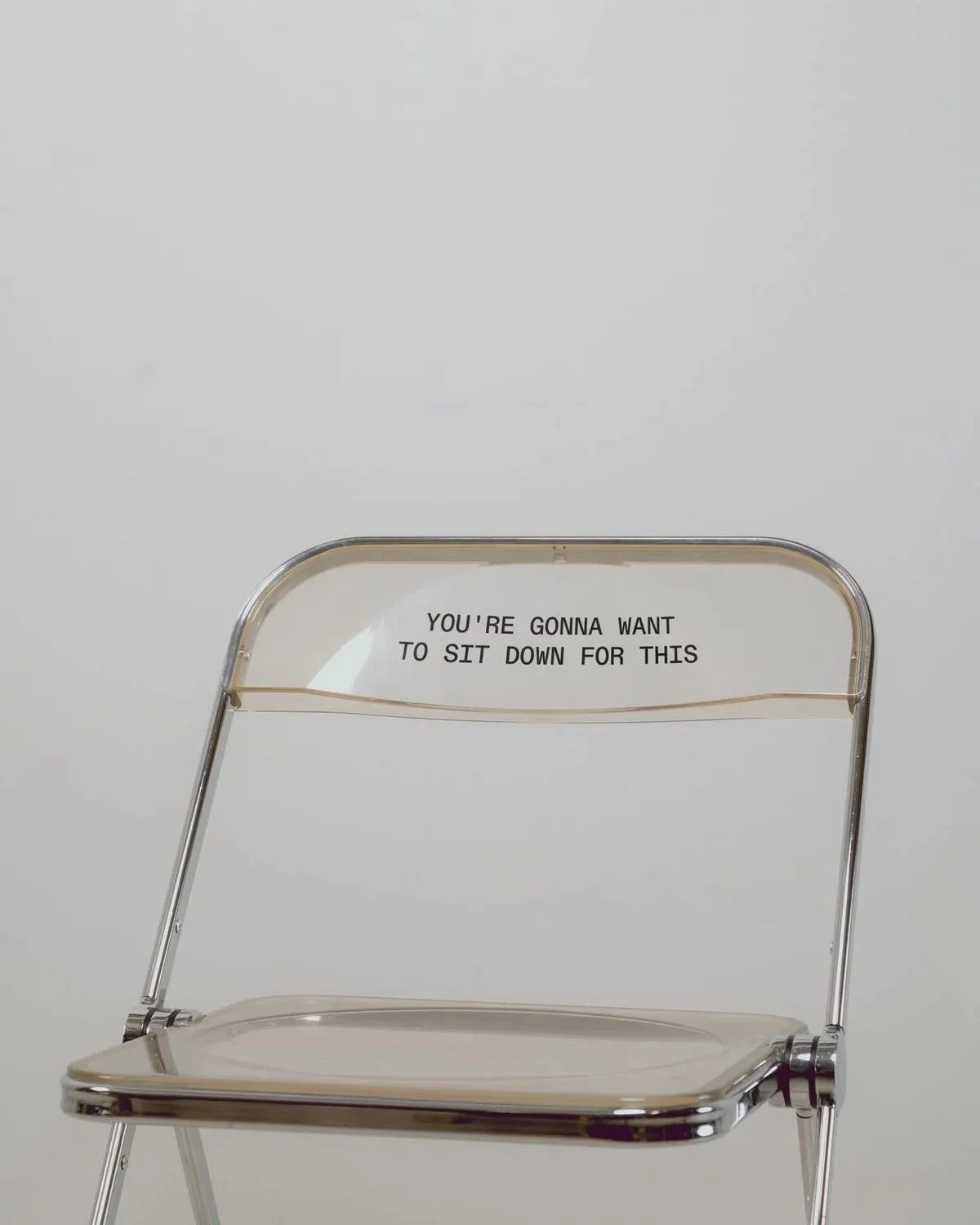A clear acrylic folding chair with a sign attached to the backrest that reads, "YOU'RE GONNA WANT TO SIT DOWN FOR THIS."