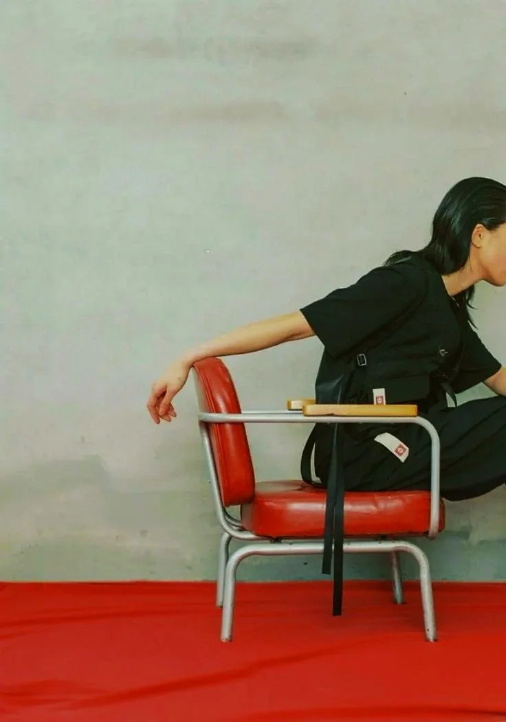 Person sitting sideways on a red school desk and chair with an arm hanging off the back of the chair.