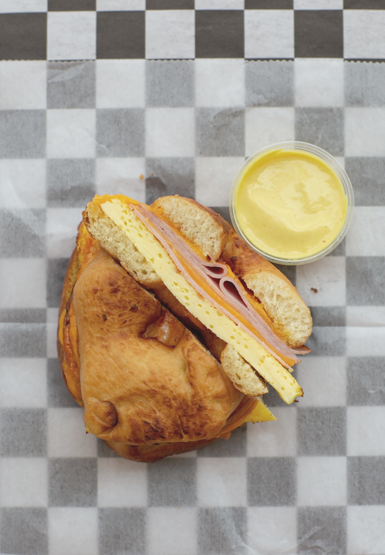 Ham and cheese sandwich on a bun with a side of egg and ham, served with a small cup of yellow mustard or sauce, on checkered paper.