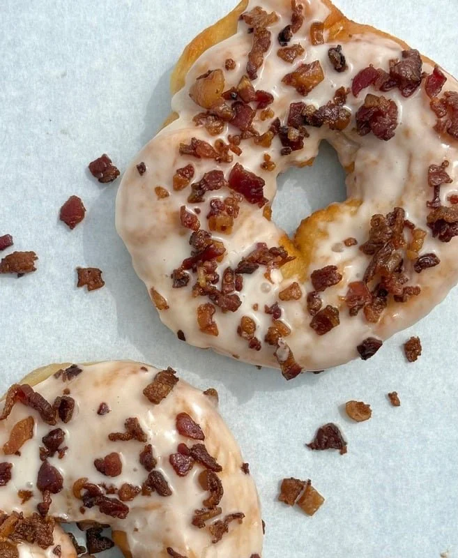 Two glazed donuts topped with bacon bits, placed on light blue paper with some bacon bits scattered around.