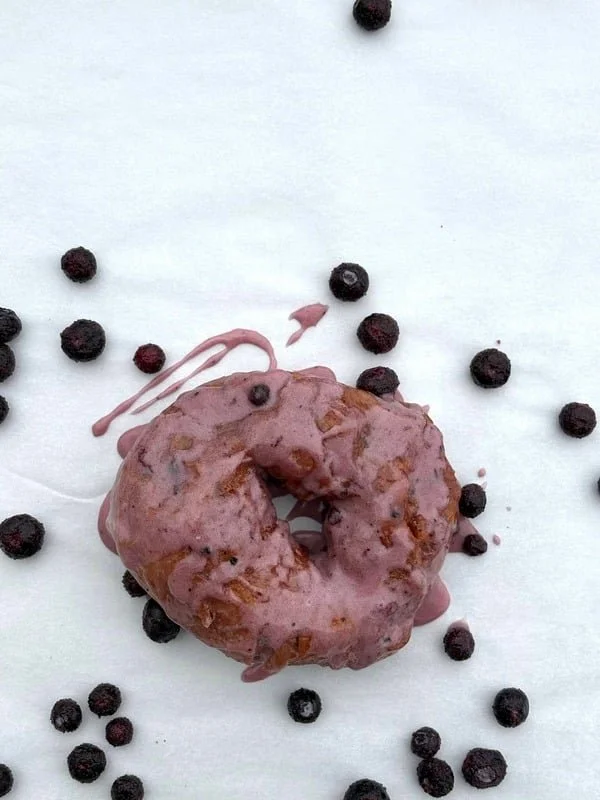 A pink glazed donut with visible nuts, surrounded by scattered blueberries on a white surface.