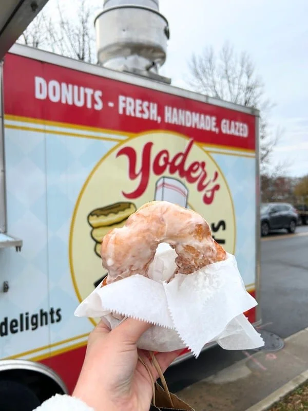 A hand holding a glazed donut with a hole in the middle, wrapped in white paper, in front of a red and white sign for Yoder's Donuts, featuring images of donuts and the words "Donuts - Fresh, Handmade, Glazed".