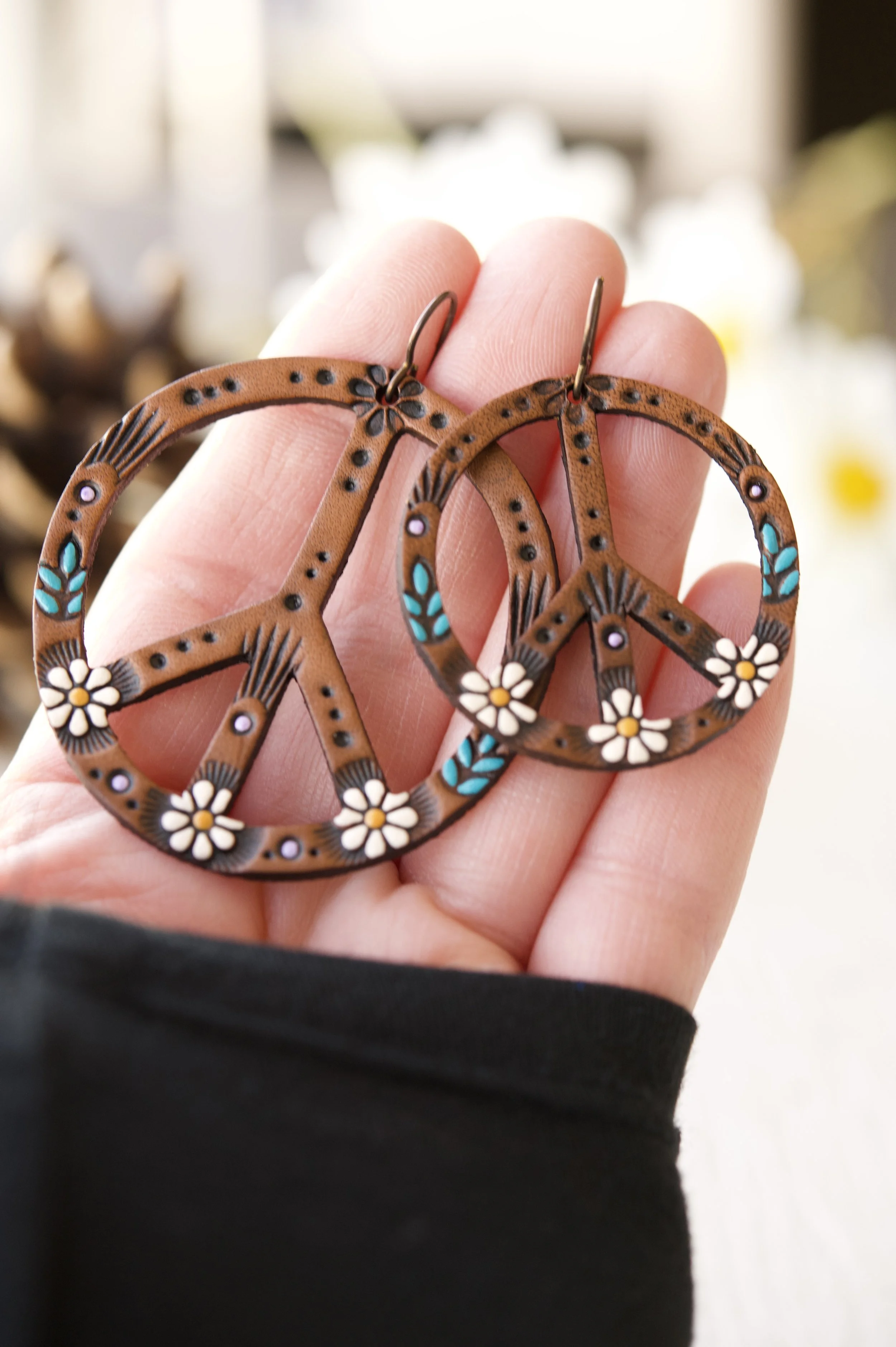 this photo shows one each of the 1.5” and 2” daisy peace sign earrings held in Brandy’s hand, the maker and owner of Mesa Dreams Leather