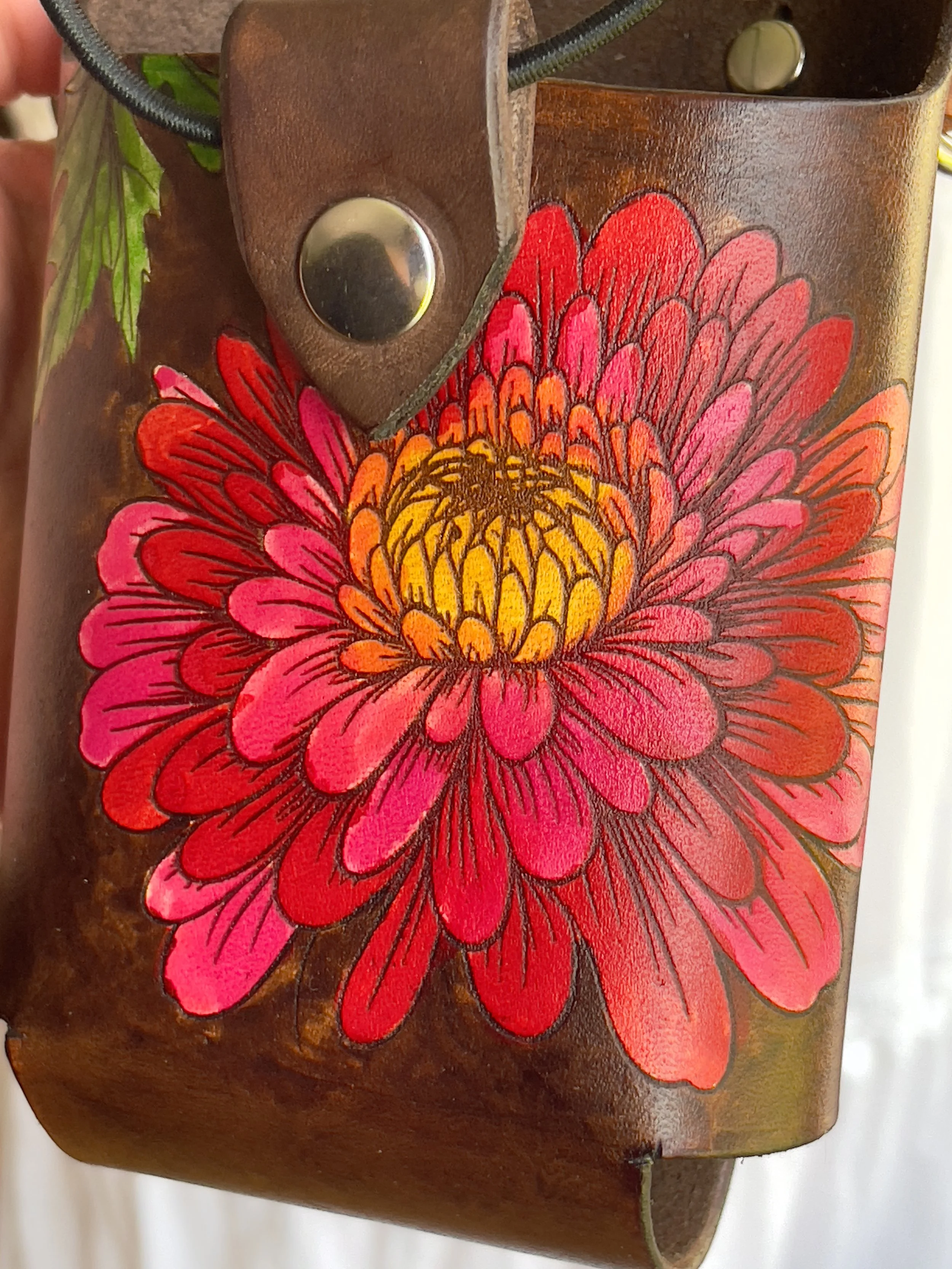 a close-up of a brown leather fire radio bucket with an engraved chrysanthemum design, painted in vibrant colors with a silver snapped tab and elastic retention cord.