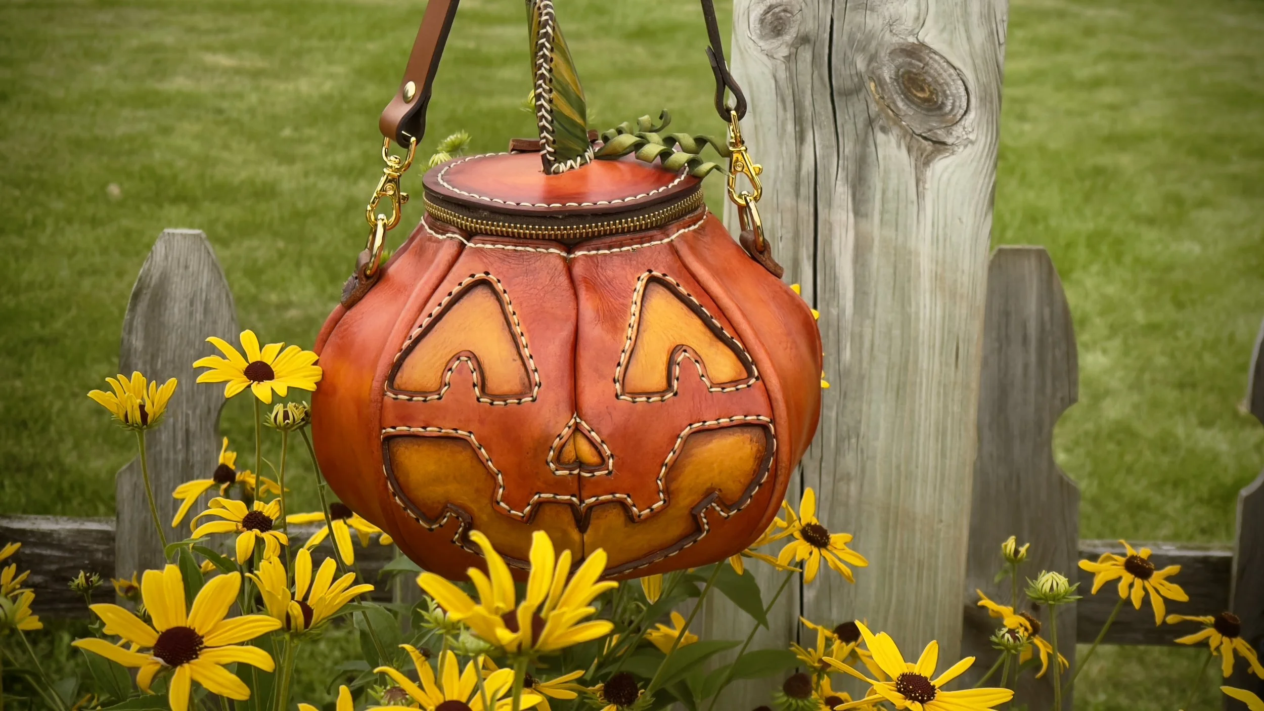 A detailed, handmade leather bag carved to resemble an orange Jack-O-Lantern hangs in a grassy meadow surrounded by Black-Eyed Susans. The unique bag features a handstitched face and green variegated stem with curly tendrils.