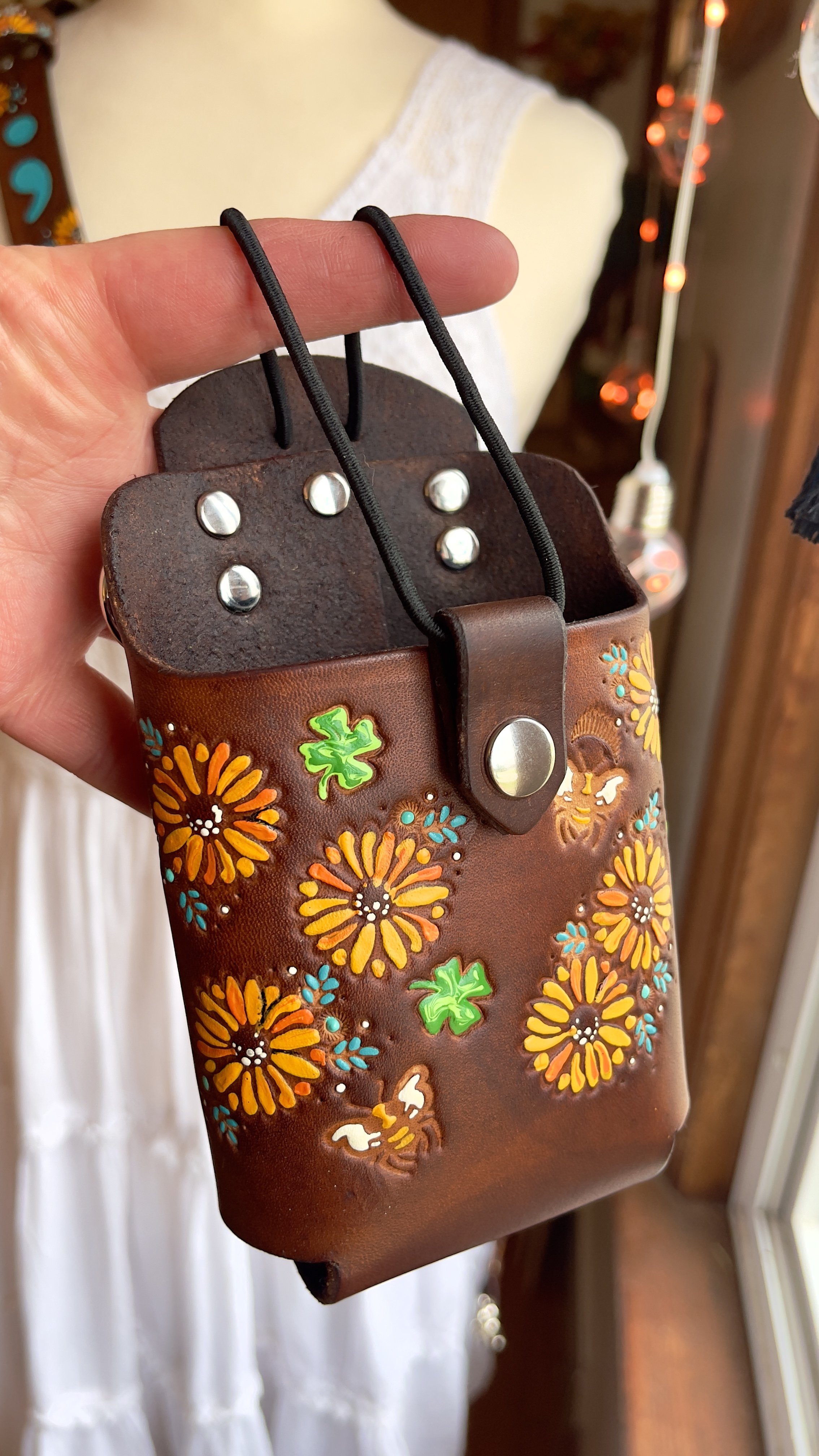 Hand-tooled brown leather radio holster with hand-painted sunflowers, green clovers, and honeybees. Features silver rivets, a snap closure, and a black elastic cord for radio security.