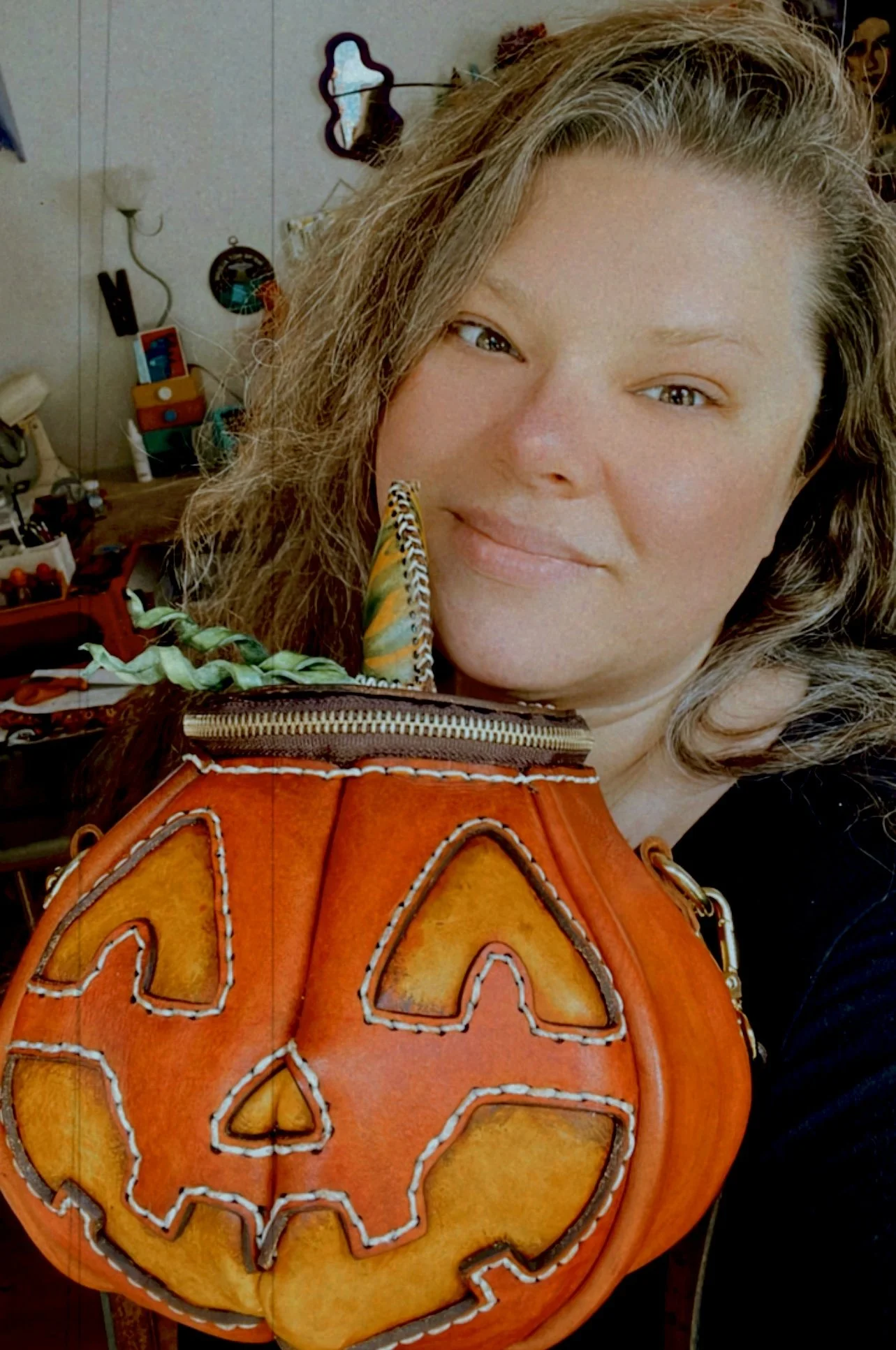 Brandy, the owner of Mesa Dreams, smiles while holding a handcrafted leather jack-o'-lantern purse. The bag features vibrant orange leather, intricate white hand-stitching, and a zipper closure at the top with green leather vine accents.