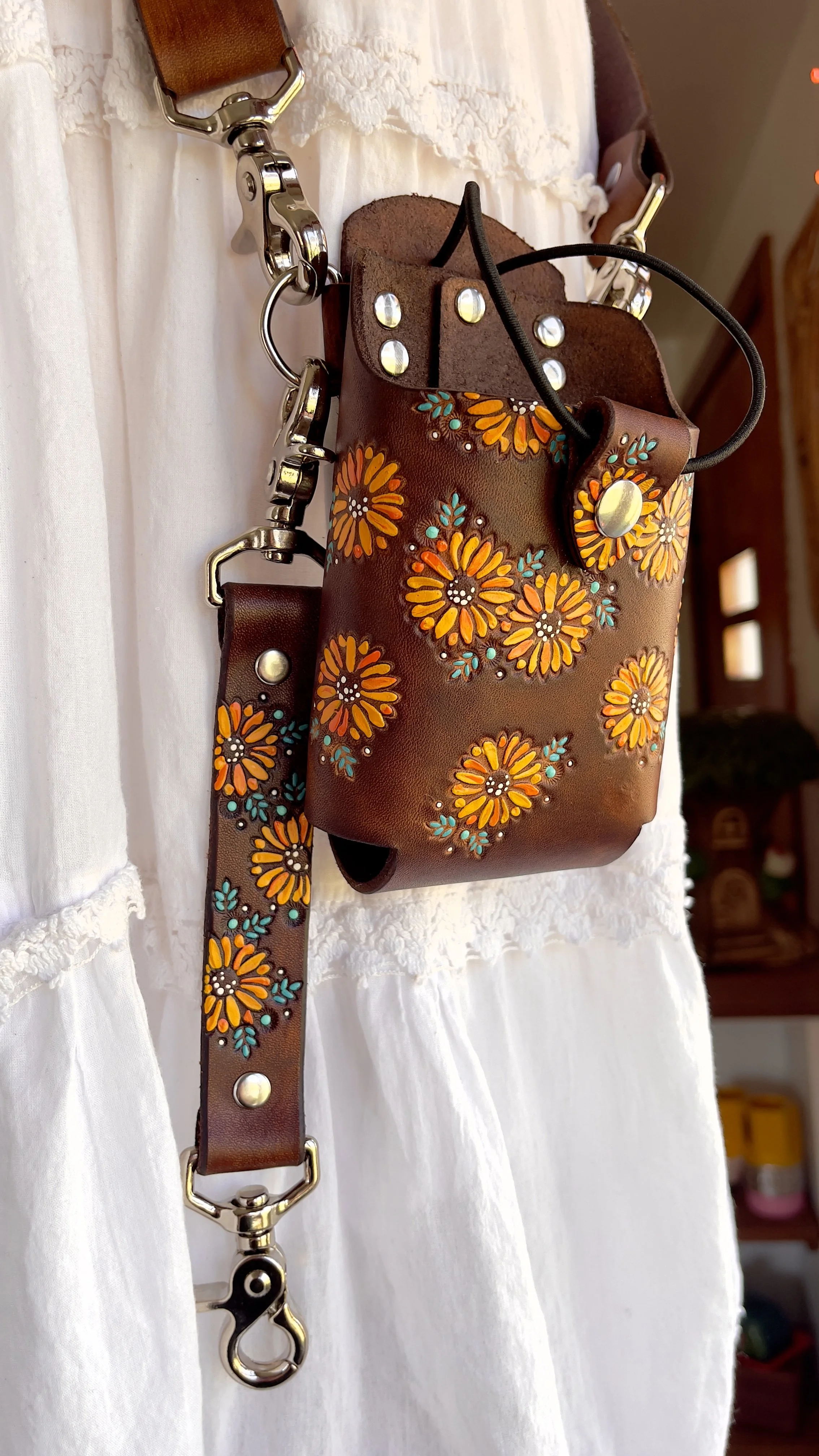 a brown leather fire radio bucket covered in hand tooled yellow sunflowers and turquoise leaves. It has a silver snap tab and black elastic retaining loop.