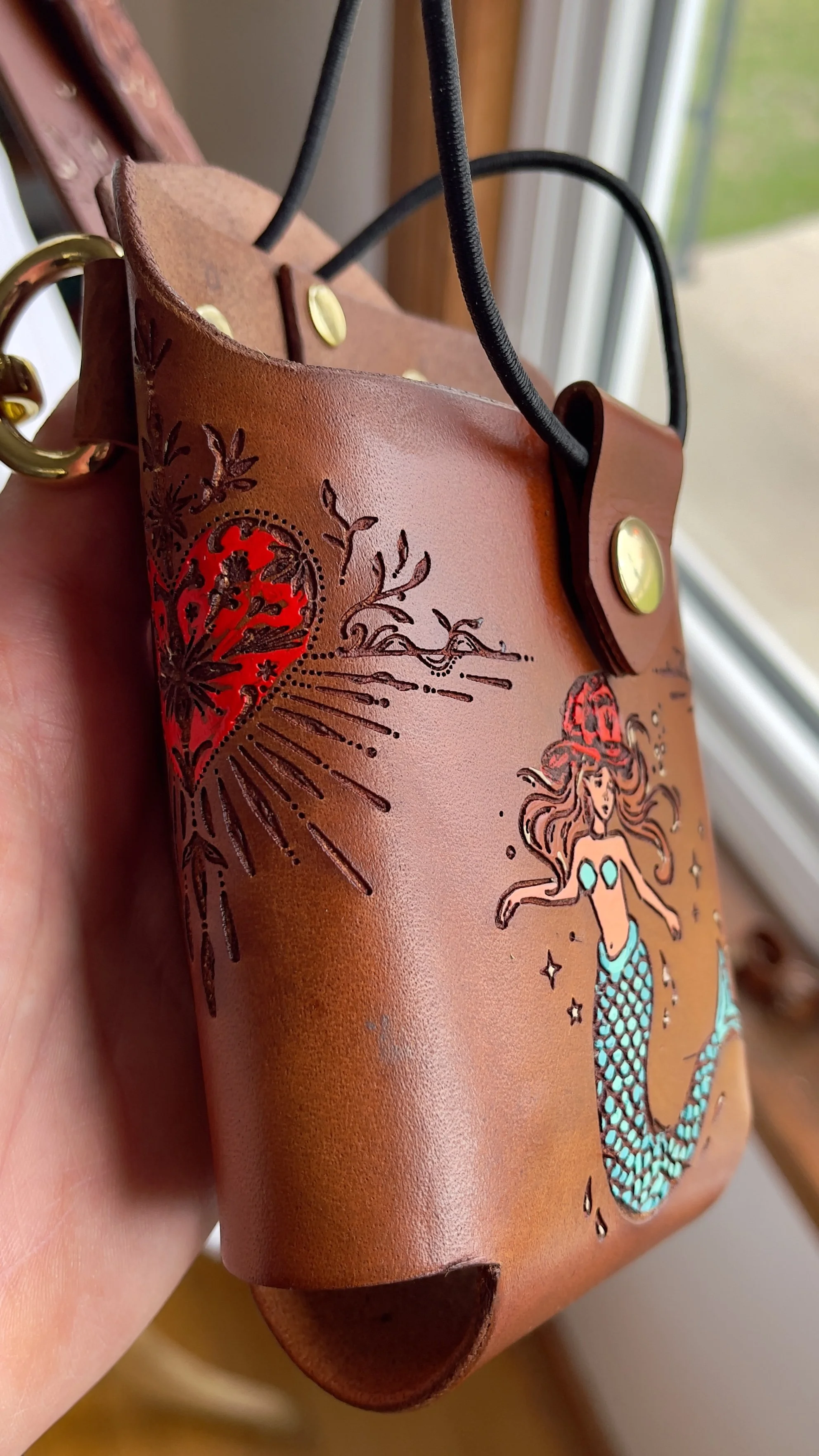 a brown leather fire radio holster engraved with a sacred heart and a mermaid wearing a fireman’s hat, both are hand painted in vibrant colors