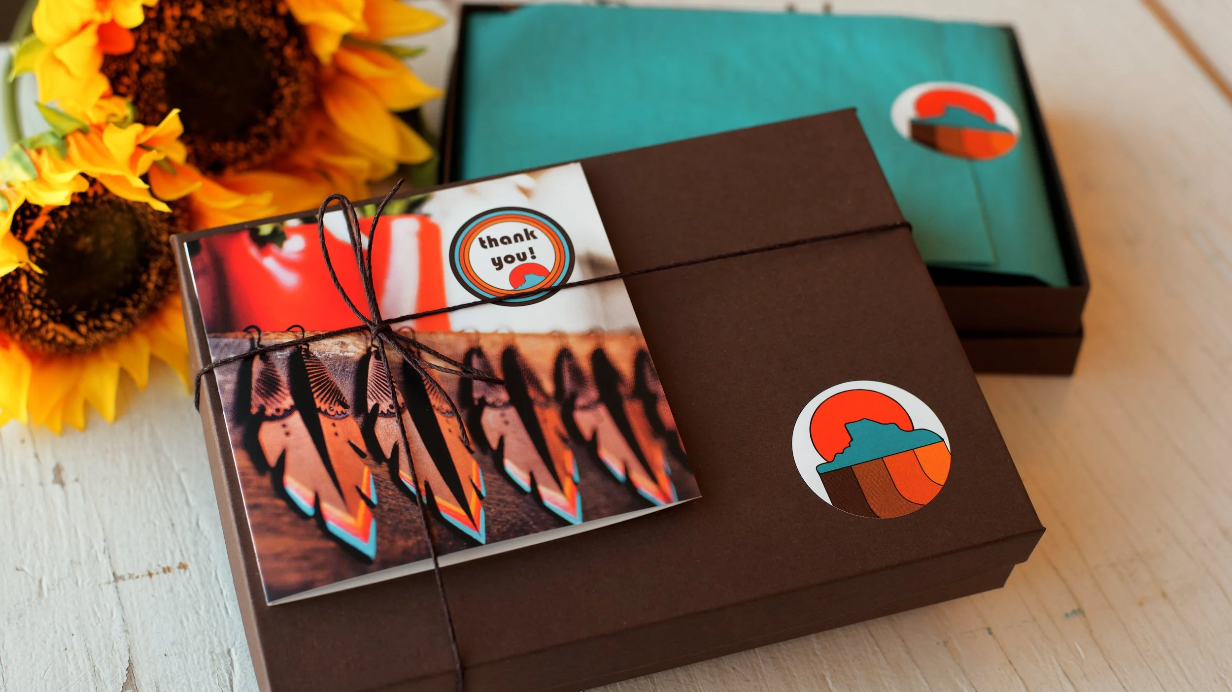 the image shows our signature chocolate brown gift boxes with colorful thank you card on top. It’s tied with twine and shows the teal tissue paper inside. It’s set against a bouquet of sunflowers on a table