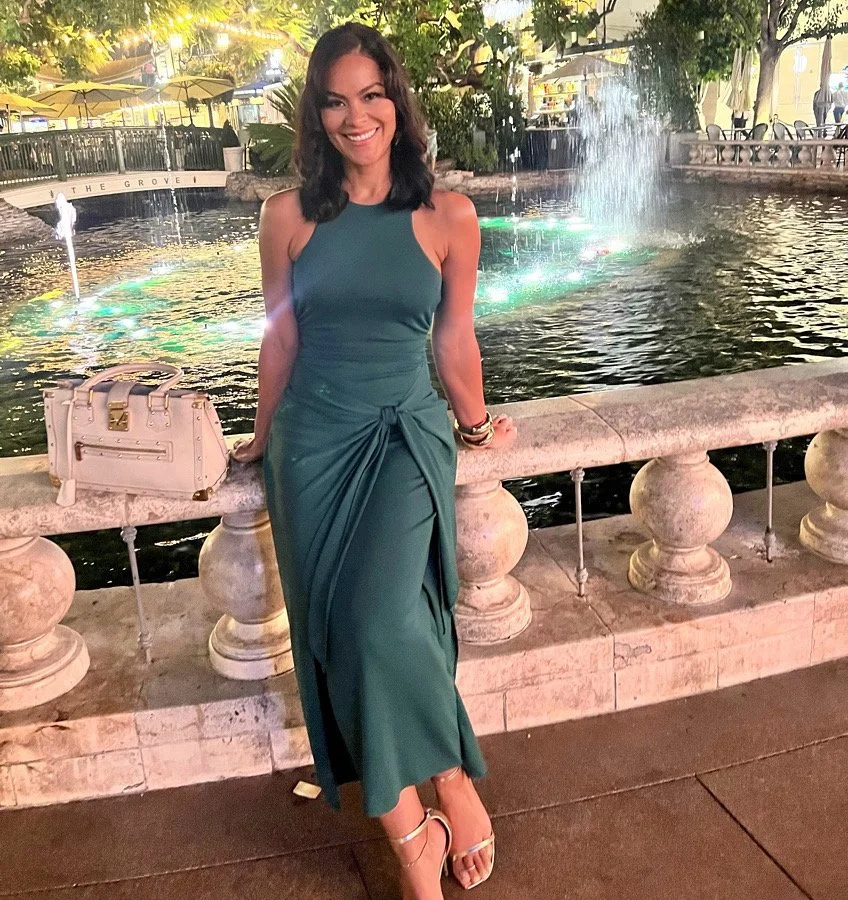 A woman with dark wavy hair wearing a teal dress standing beside a stone railing next to a fountain in a decorative outdoor setting at night.