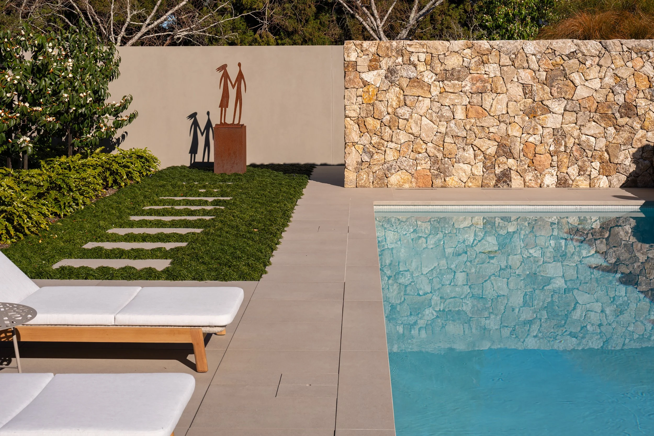 A modern backyard scene featuring a swimming pool with clear blue water, a white lounge chair, a stepping stone pathway through a lawn, a stone wall, a beige wall with a metal sculpture of a couple holding hands, and lush greenery including small trees and bushes.