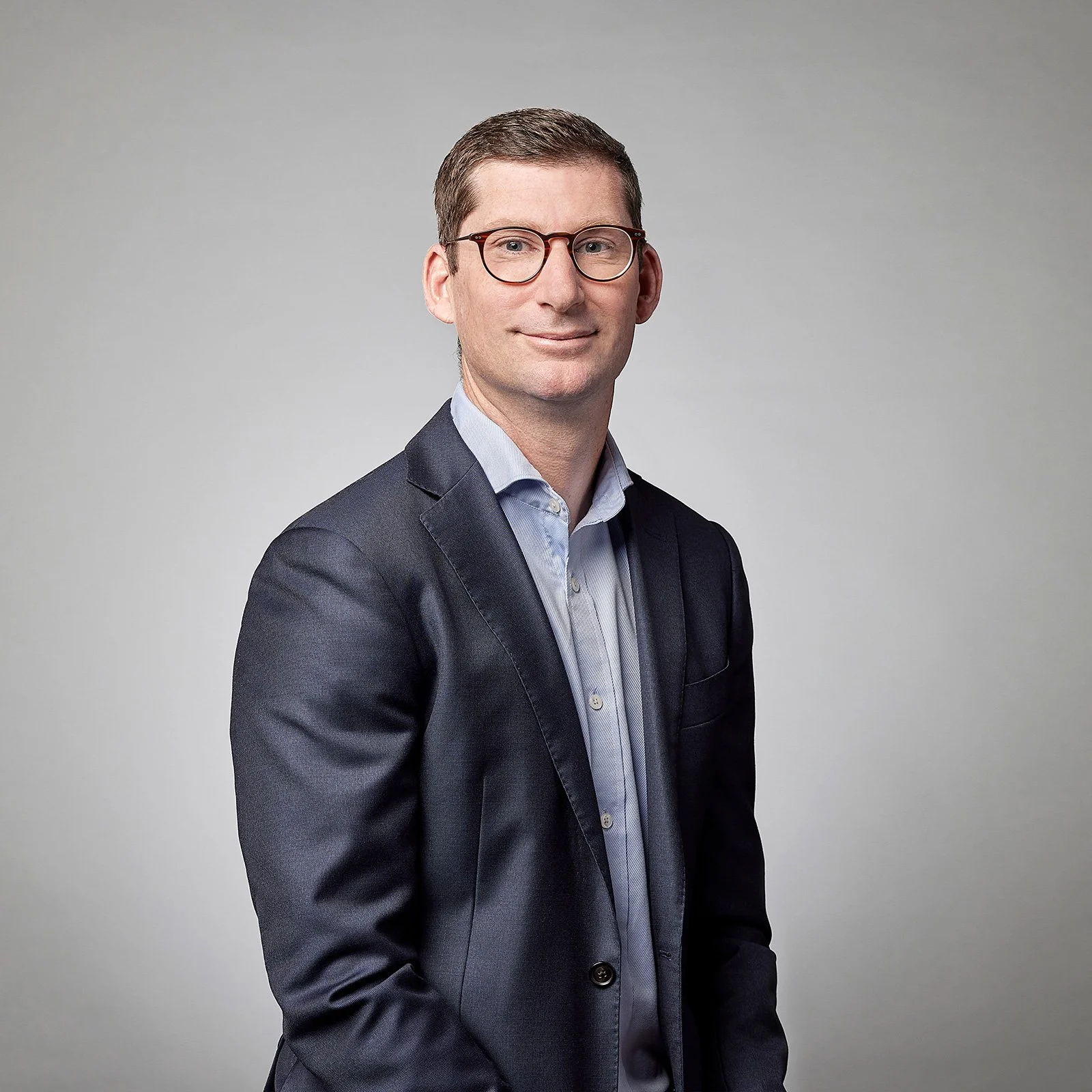 A man in a dark suit and light blue shirt with glasses, smiling at the camera against a grey background.