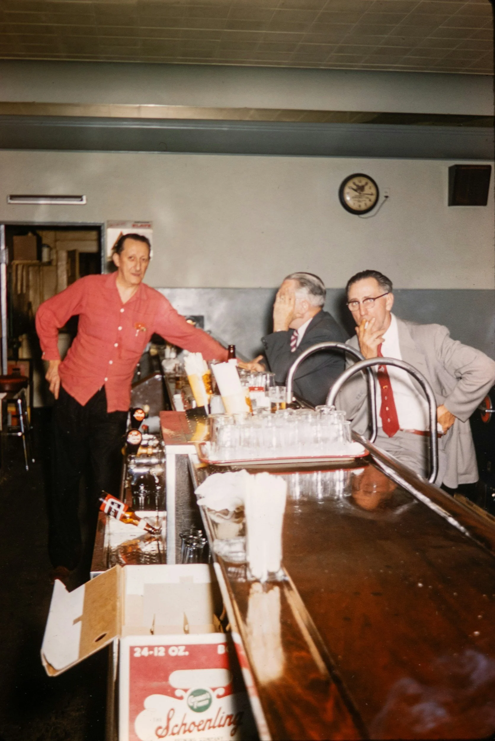 A bar setting with three men sitting and a woman bartender standing behind the counter. The men are dressed in suits and tie-like attire, one is holding his face with his hand, and another is smoking a cigarette. The bartender is wearing a red shirt and is reaching for something on the counter. The bar has various bottles and glasses, and there is a clock on the wall showing the time as approximately 9:10.