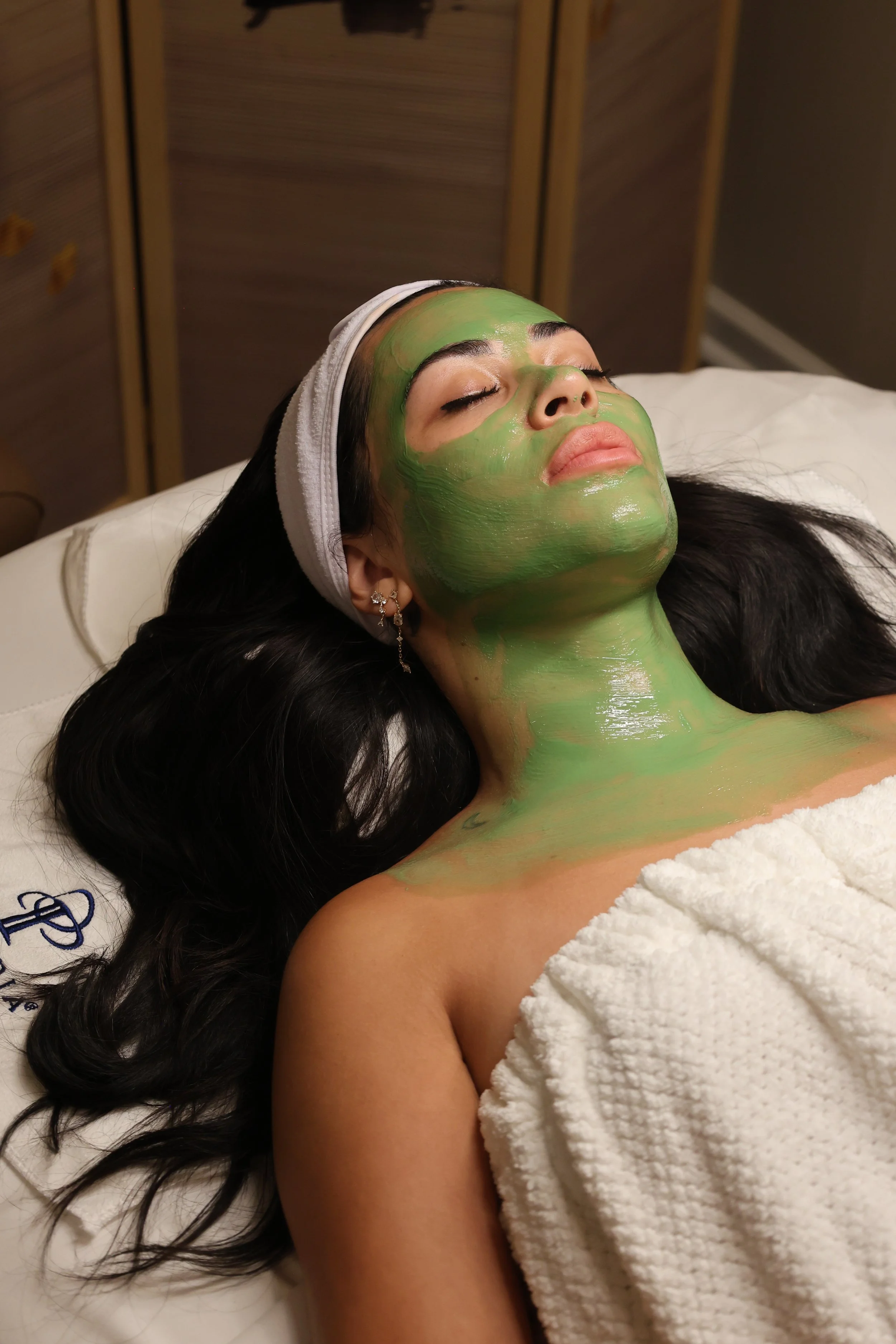 A woman with dark hair lying on a bed with a towel wrapped around her chest, receiving a green facial mask treatment in a spa or salon setting.