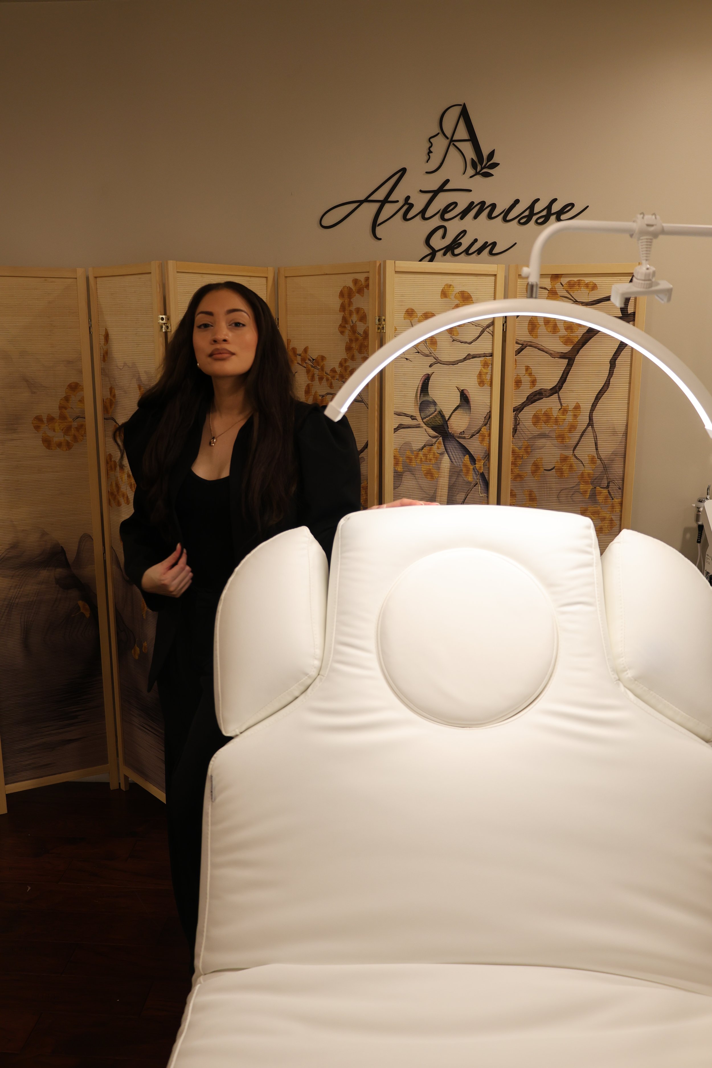 A woman with long dark hair wearing a black outfit standing in front of a decorative folding screen with golden floral and bird designs. Behind her is a wall with the sign 'Artemisse Skin'.