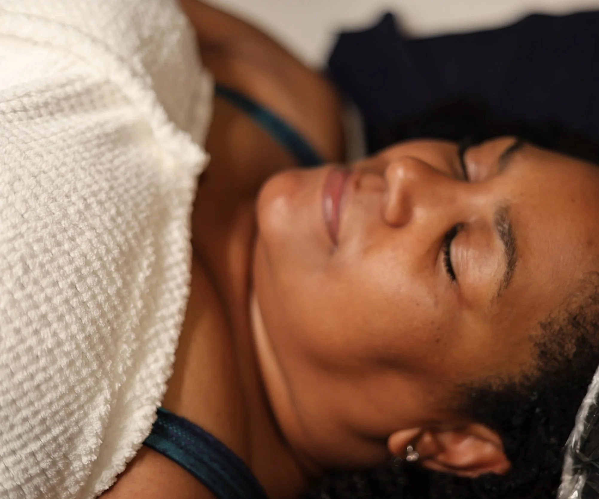 Close-up of a woman's face with her eyes closed, lying down with a beige towel covering part of her face, and a dark shirt visible.